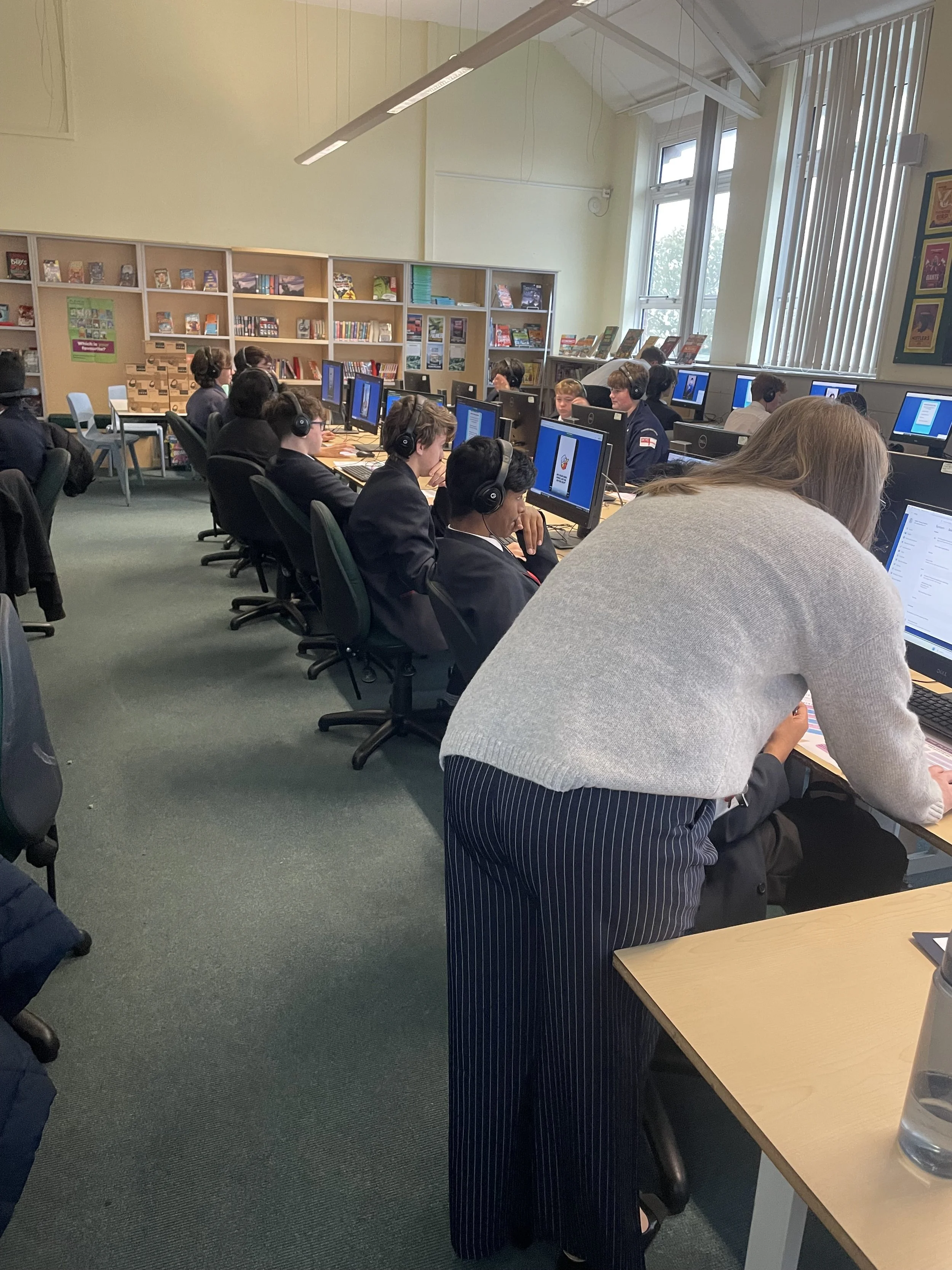 Students working on computers in a classroom with bookshelves, windows, and vertical blinds.
