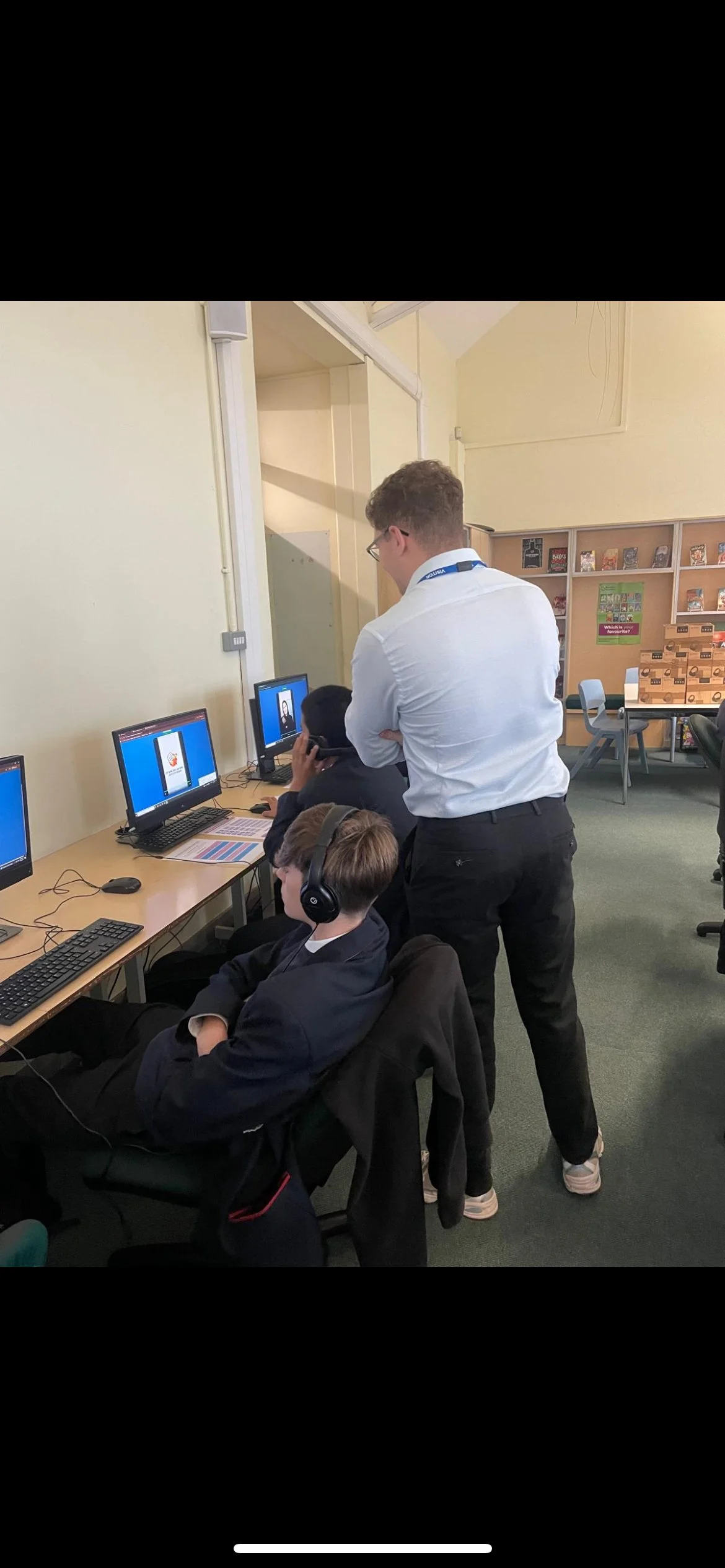 Two boys in school uniforms at computer workstations, with a teacher supervising in a classroom that has beige walls, computers, and educational materials.