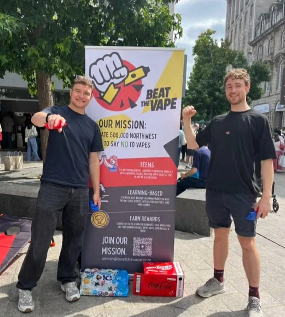 Two young men standing outdoors in front of a colorful banner promoting a campaign called 'Beat the Vape.' They are smiling and flexing their right arms. The banner includes a fist holding a vape device, with the mission to prevent 500,000 northwest teens from vaping. There are boxes of soda and snacks on the ground in front of them.