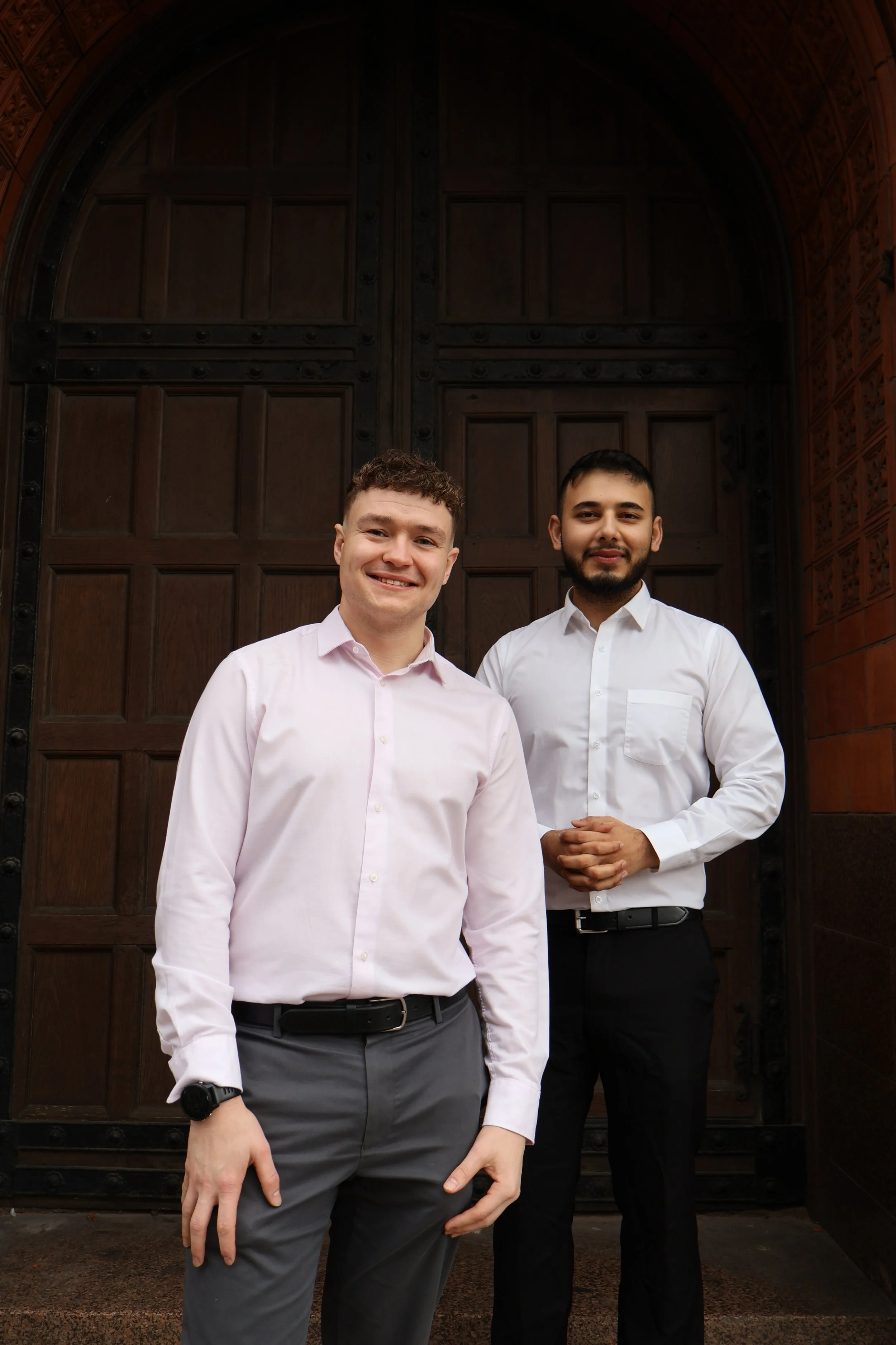 Two young men standing in front of a large wooden door, dressed in business casual attire, smiling.