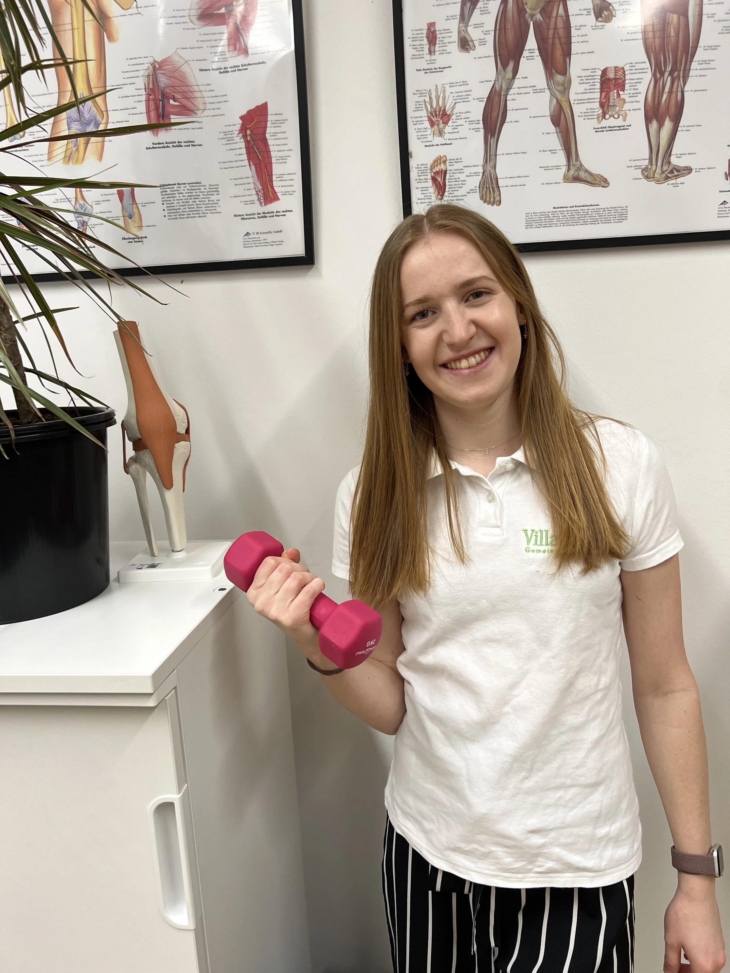 Junge Frau hält pinken Hantelarm im Fitnessstudio, im Hintergrund Anatomie-Poster des menschlichen Körpers.