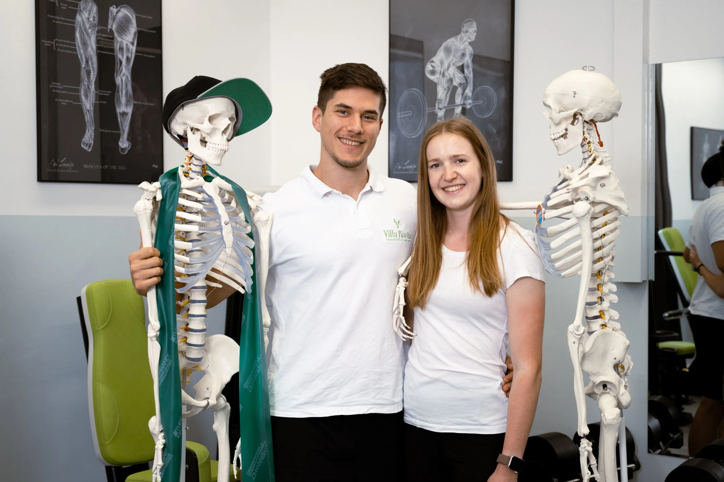 Zwei junge Menschen, ein Mann und eine Frau, stehen zwischen zwei menschlichen Skeletten in einem medizinischen Unterrichtsraum. Hinter ihnen sind Anatomie-Poster an der Wand zu sehen.