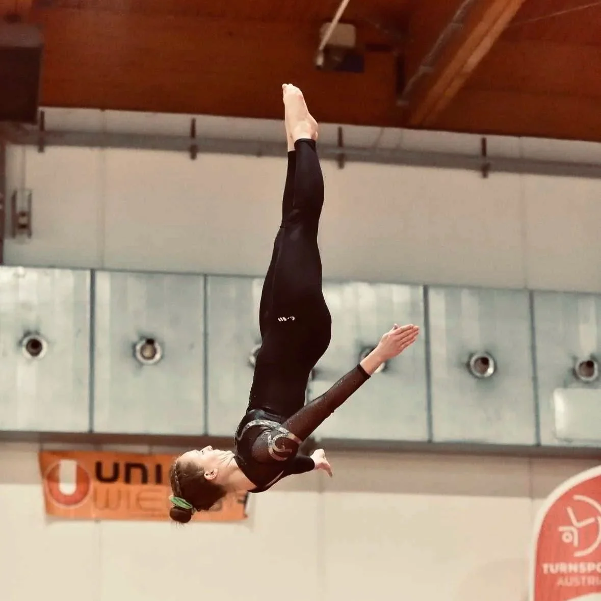 Gymnastin führt eine Sprung- oder Drehübung auf einer Turnmatte im Turnhallenraum aus.