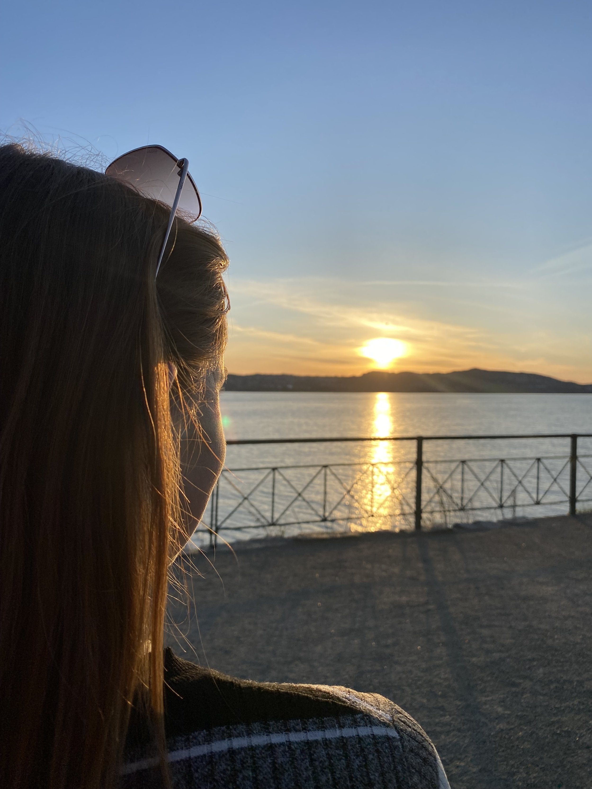 Eine Person mit braunen Haaren und Sonnenbrille schaut auf einen Sonnenuntergang über einem Fluss oder See, im Hintergrund Berge und ein Himmel mit blauer Farbe und warmen Tönen.