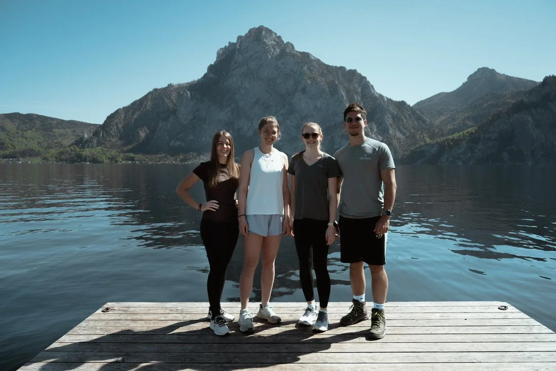 Vier junge Menschen stehen auf einem Steg vor einem See mit Bergen im Hintergrund, sonniger Tag, entspannte Atmosphäre.