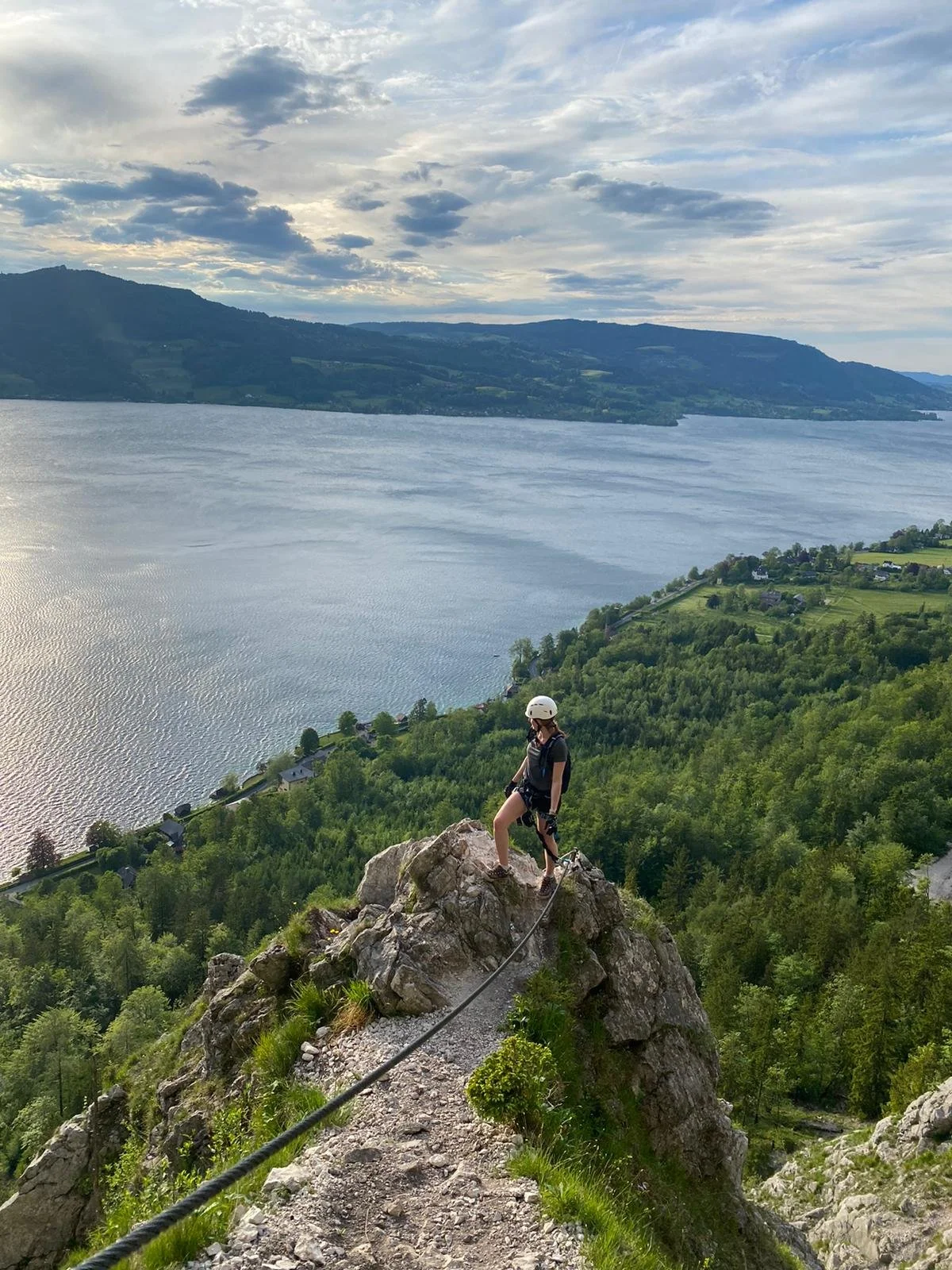 Kletterer auf einem Felsen mit Blick auf einen See, grüne Landschaft und Berge in der Ferne.
