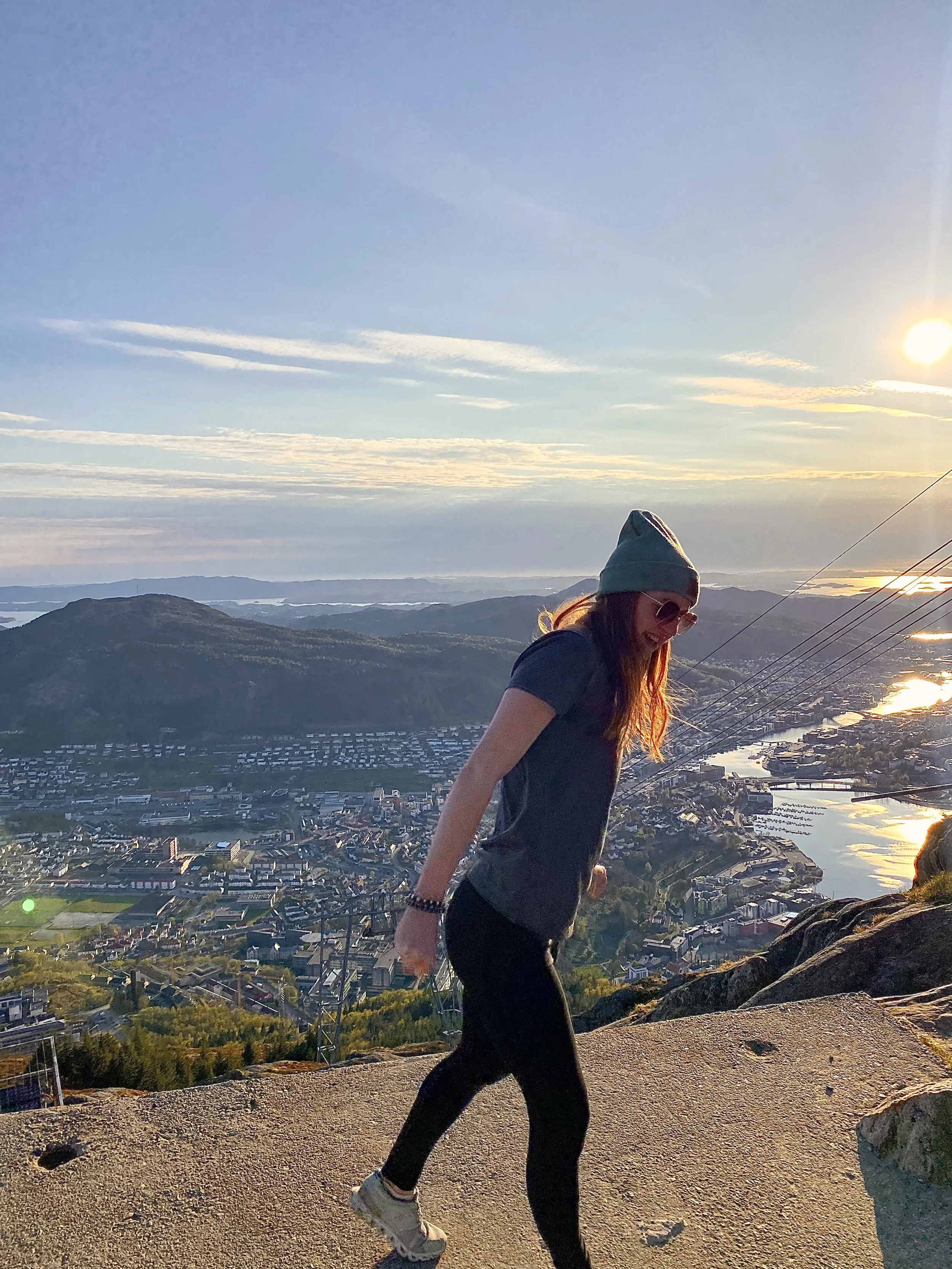 Eine junge Frau trägt eine graue Mütze, eine Sonnenbrille, ein schwarzes T-Shirt und schwarze Leggings, während sie auf einem Berggipfel bei Sonnenuntergang steht, mit Blick auf eine Stadt und einen Fluss unter einem blauen Himmel mit leichten Wolken.