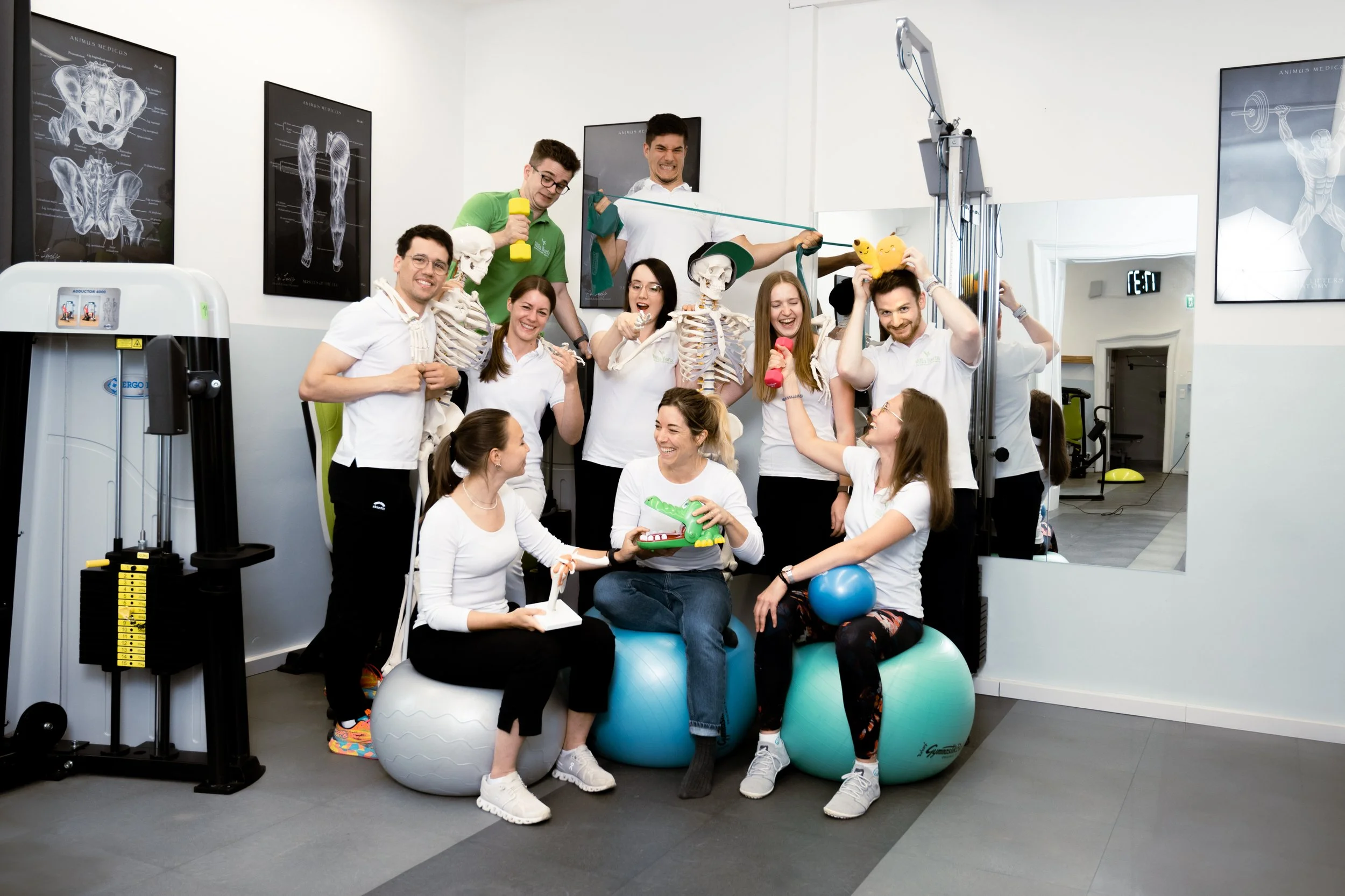 Eine Gruppe von Menschen in einem medizinischen oder physiotherapeutischen Raum, die mit Skelletmodellen und Fitnessgeräten spielerisch trainieren, lachen und Spaß haben.