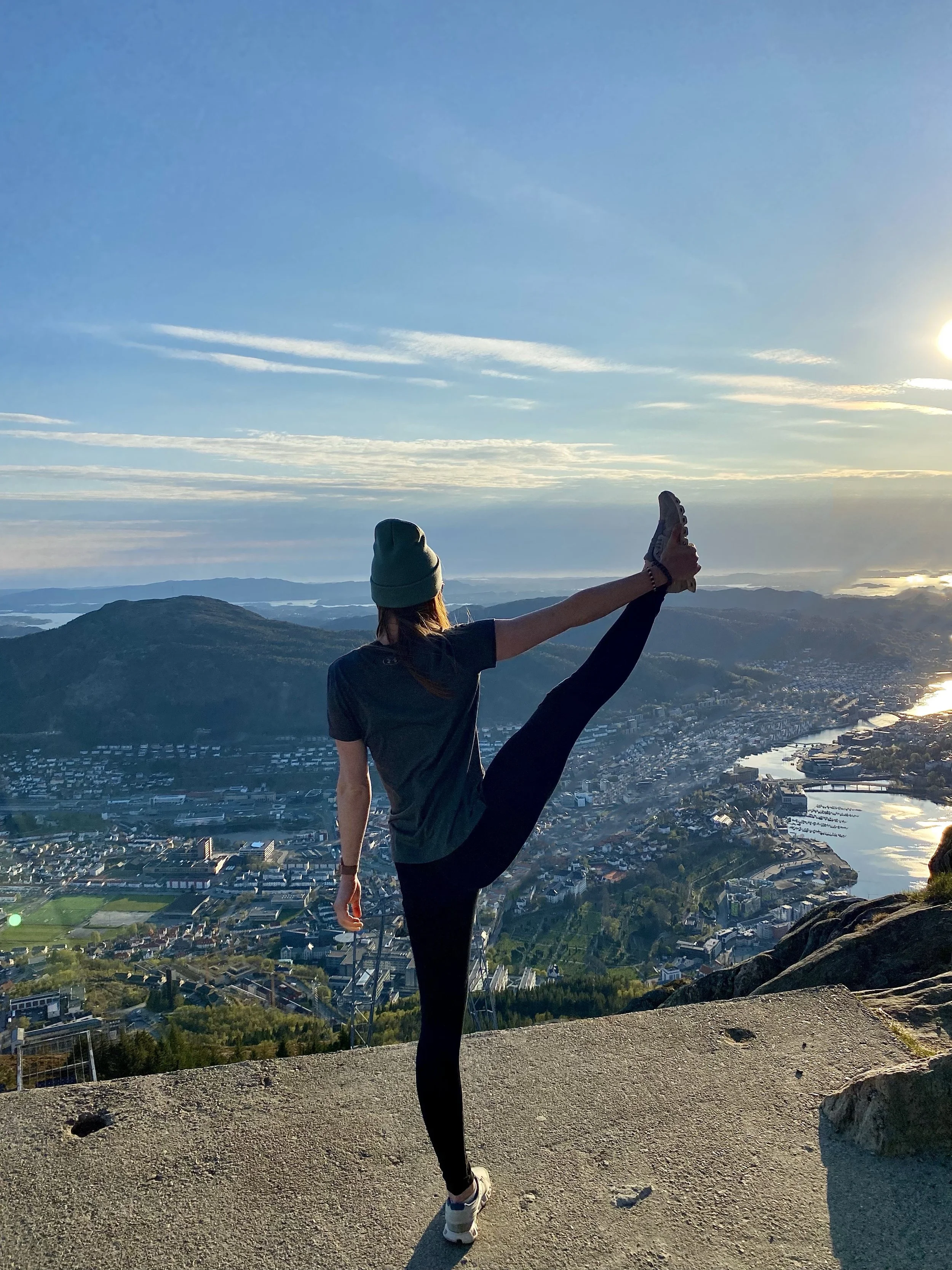 Eine junge Frau macht im Freien mit Blick auf eine Stadt, Hügel und Wasser bei Sonnenaufgang oder Sonnenuntergang einen Yoga- oder Stretching-Übung.