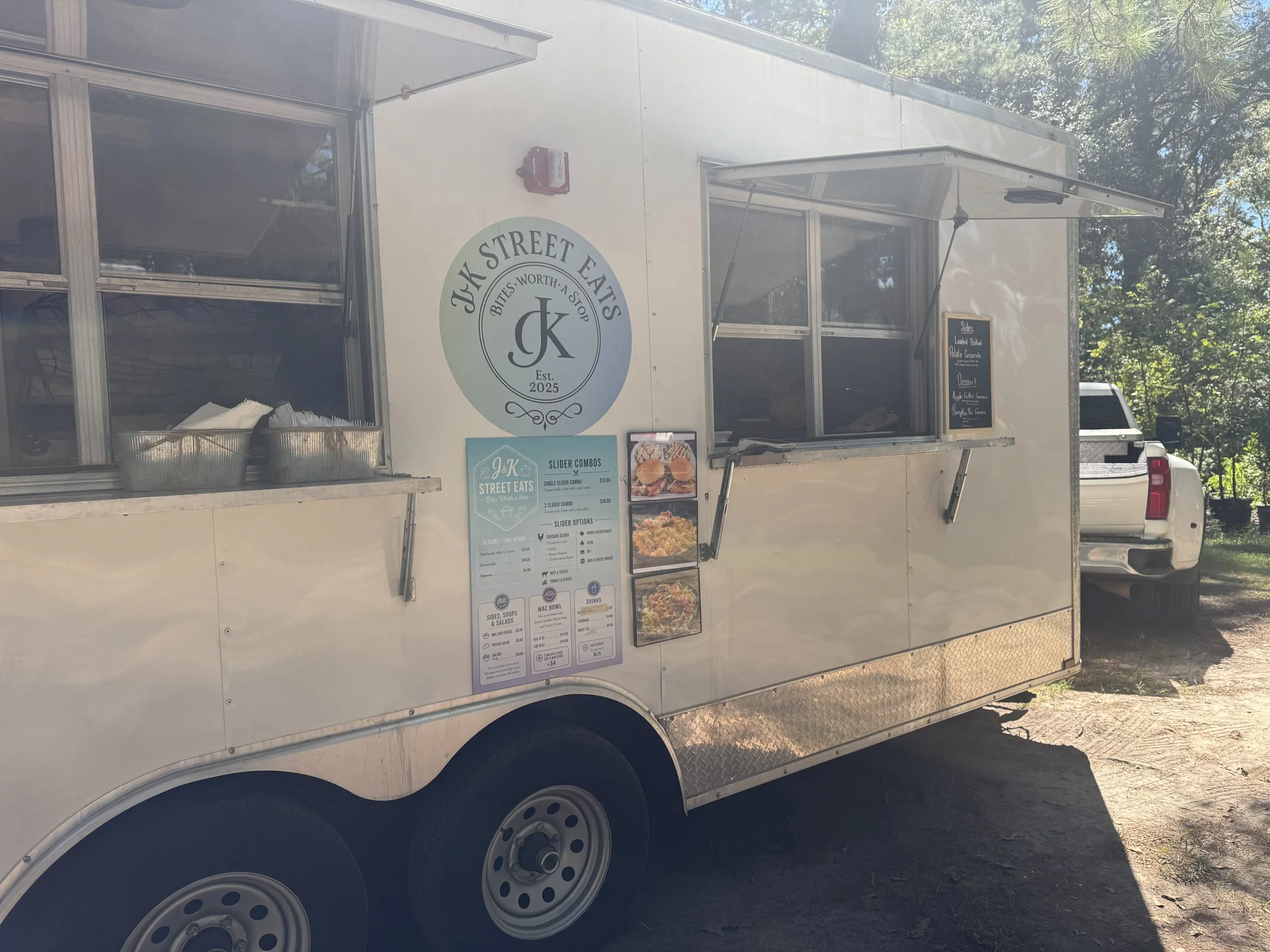 White food truck named 'JK Street Eats' with a logo that says 'Bites. Worth. A. Stop' and 'Est. 2025'. The truck has an open window with a menu sign underneath, listing sides, desserts, and drinks. The truck is parked outdoors among trees.