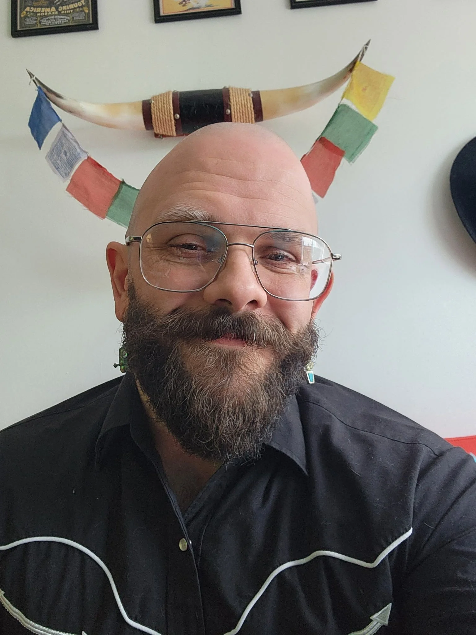 A man with glasses and a beard taking a selfie, with a decorative horn mounted on the wall behind him and framed posters above.