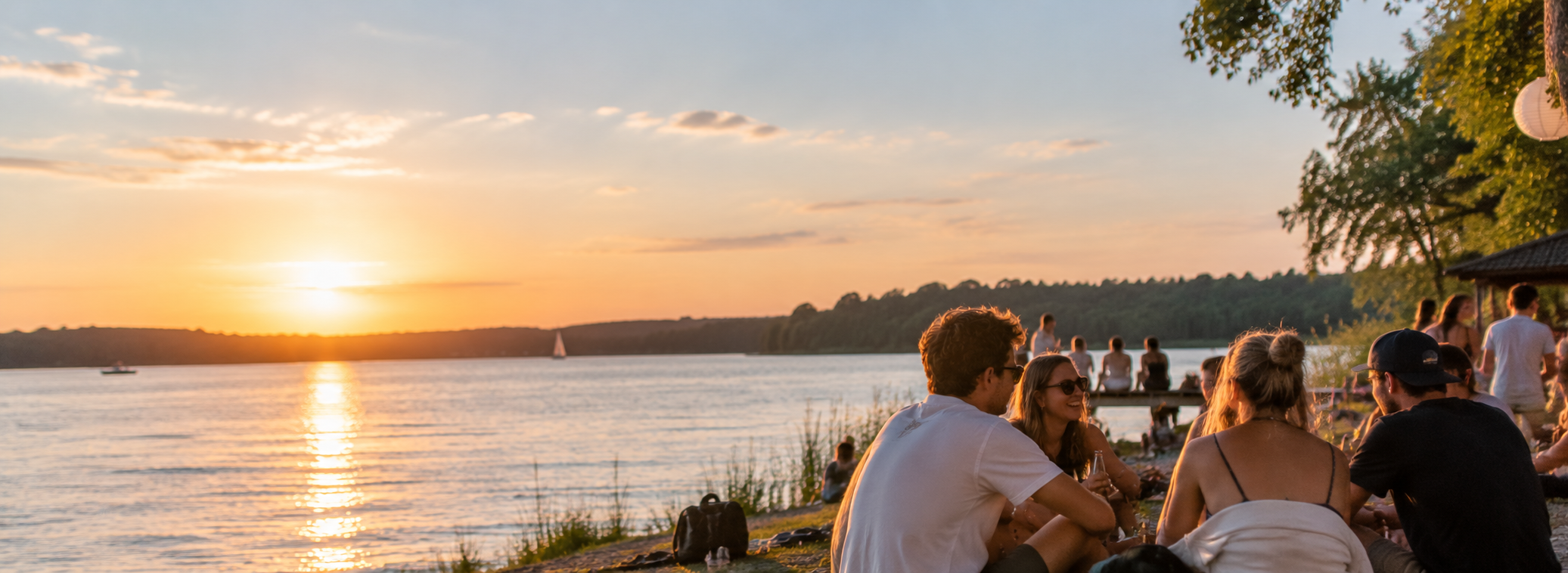 Menschen sitzen bei Sonnenuntergang am Flussufer, einige unter Bäumen, während Boote auf dem Wasser fahren.