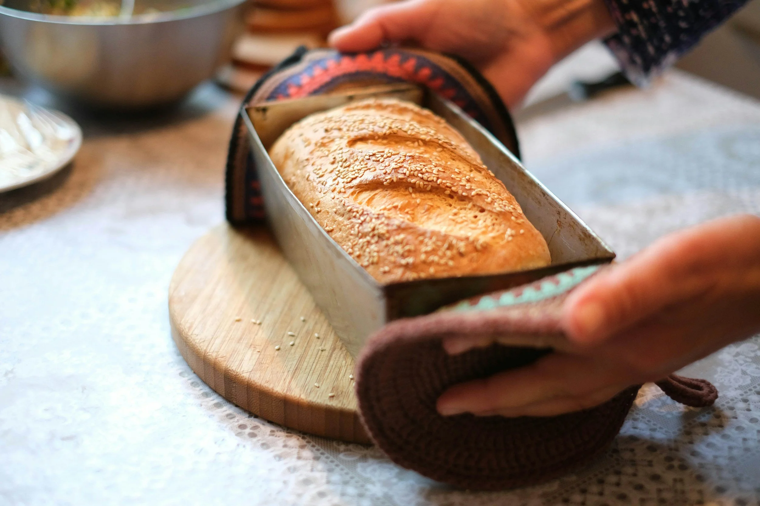 Hand pulling freshly baked bread with sesame seeds from a bread loaf tin, placed on a round wooden cutting board.