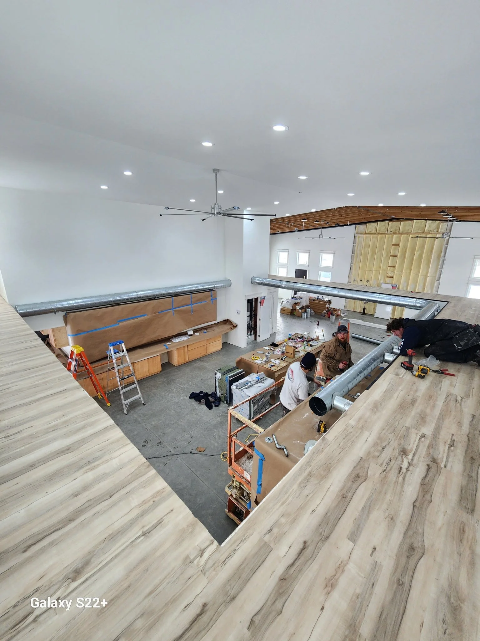 Interior of a building under construction or renovation, with workers installing ductwork and working on wooden structures, ceiling with recessed lighting, and a partially finished wall with windows.