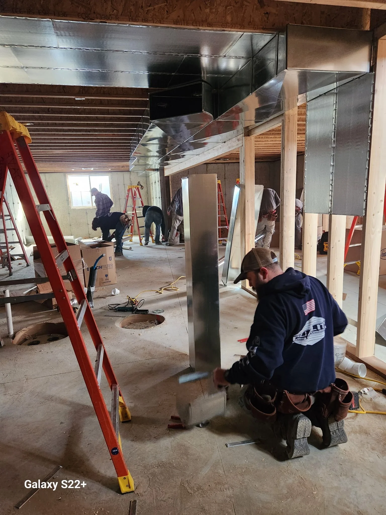 Construction workers installing ductwork in a building under construction. Several workers are working on the ceiling and walls, with ladders and tools around. Wooden support beams are visible.