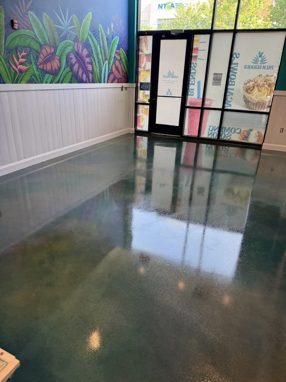 Empty interior space with a polished, shiny greenish floor, white paneled wall, colorful tropical leaf mural, and large window and door with advertisements for smoothies and desserts.