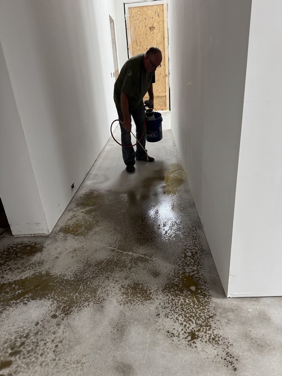 A man is using a sprayer to clean or treat a concrete floor in a hallway, with white walls and a wooden door at the end.