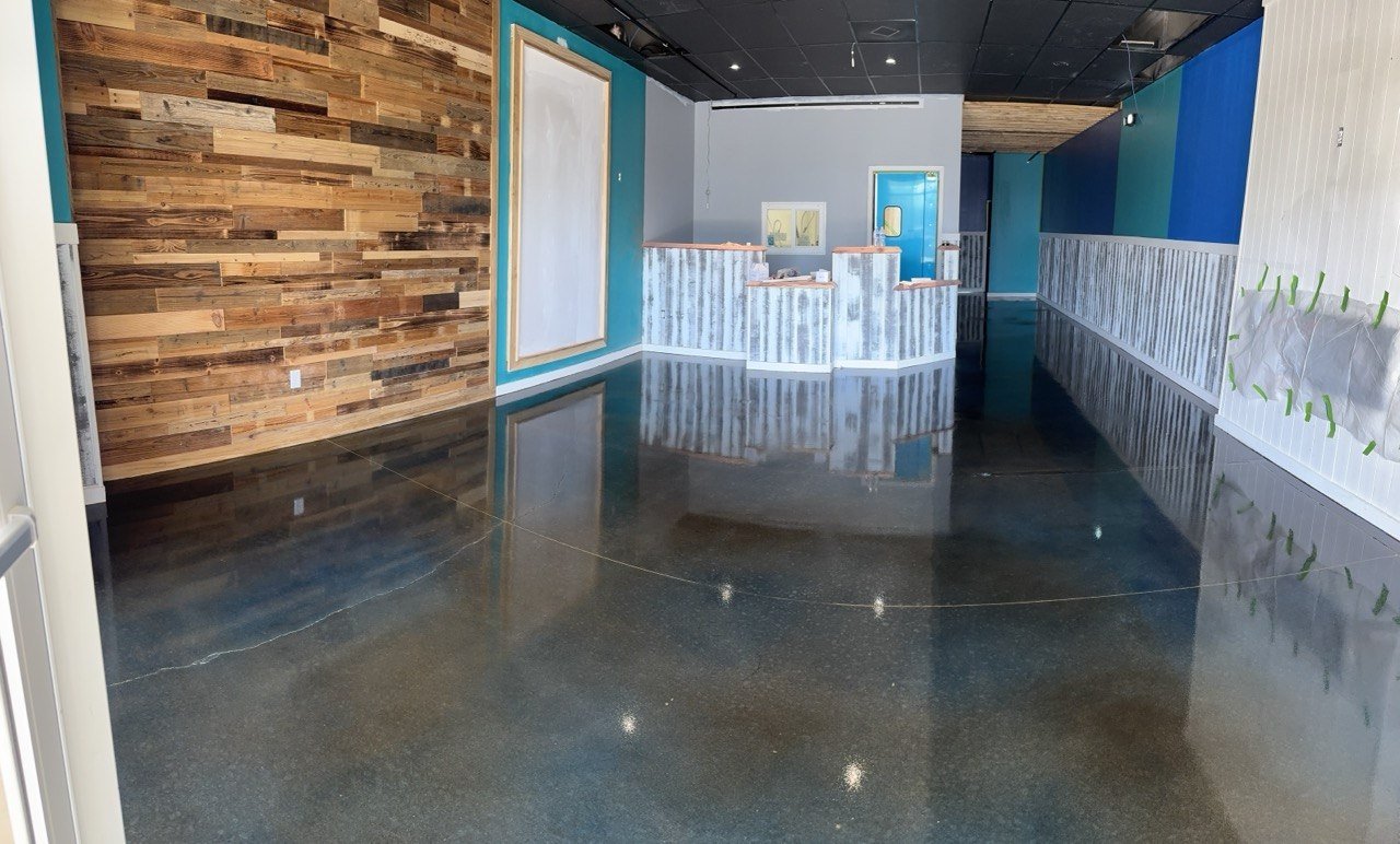 Lobby area under renovation with polished concrete floor, wooden wall on the left, painted walls in shades of blue, a white grayish reception desk at the center, and construction materials on the right.