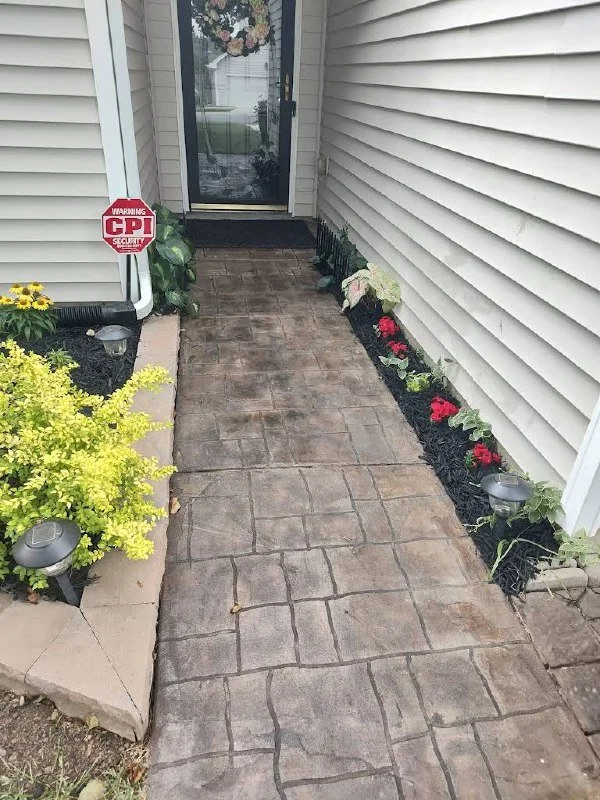 Front walkway leading to a house door, bordered by flower beds with yellow and red flowers, illuminated by small pathway lights, with a security sign on the left side.