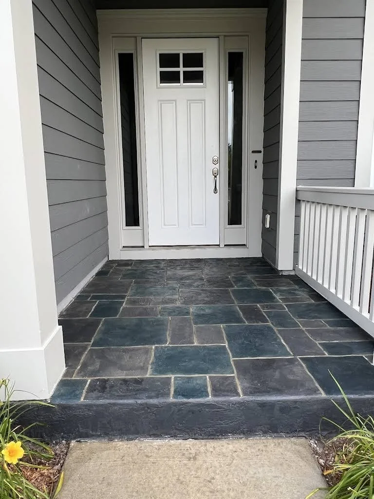 Front porch with a white door, narrow side windows, grey siding, black slate floor, and a small garden with yellow flowers.