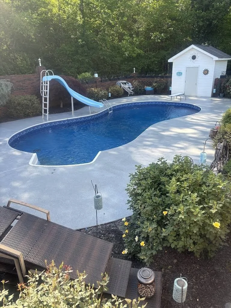 Residential backyard with a swimming pool, blue slide, poolside chairs, small white shed, bushes with yellow flowers, and outdoor lighting at dusk.