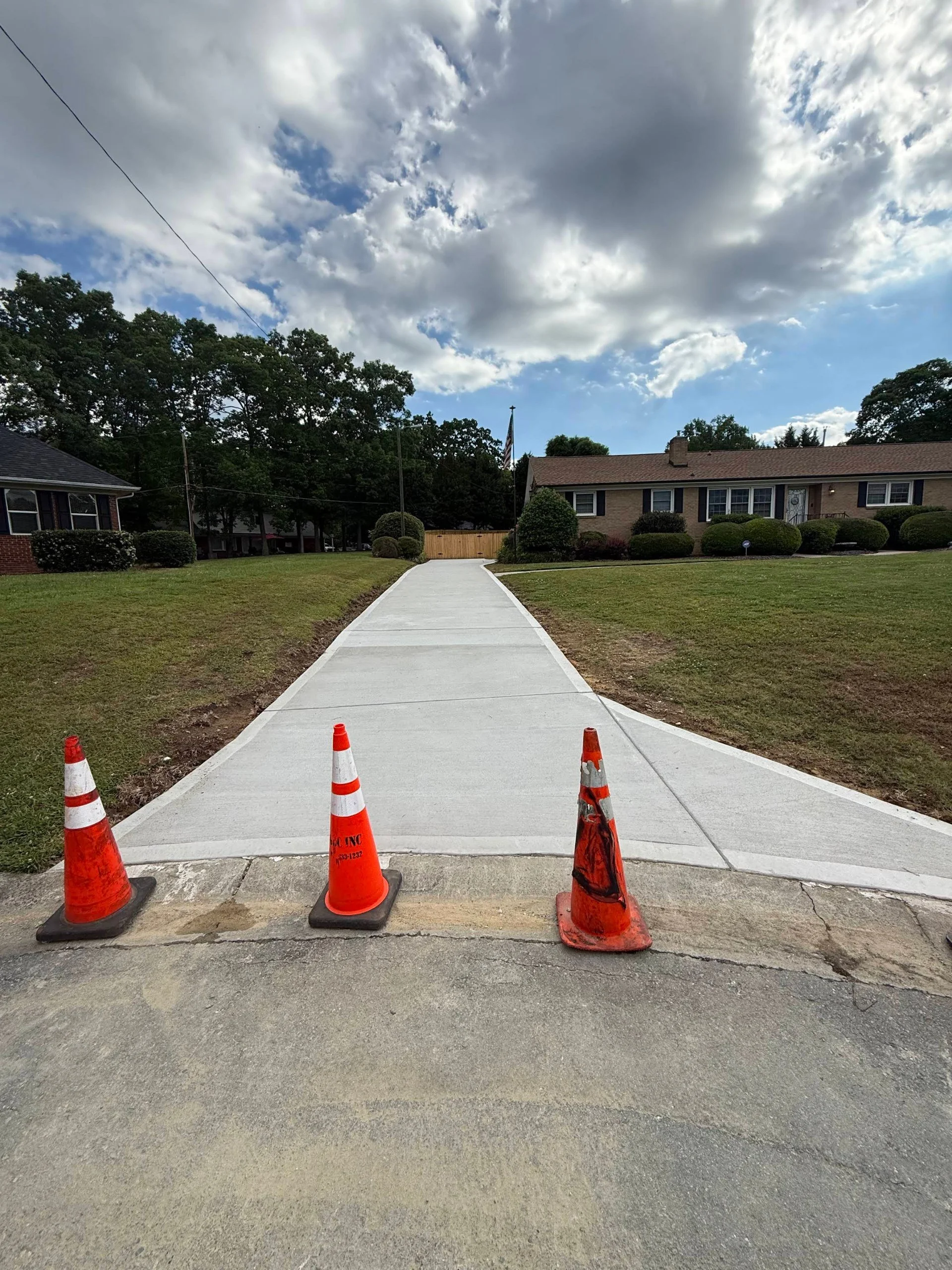 Newly paved sidewalk blocking driveway with orange traffic cones at the entrance.