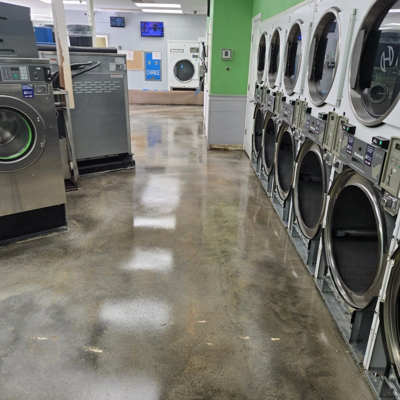 A laundromat with front-loading washing machines on the right side and dryers on the left side, with a shiny wet floor and a green wall in the background.