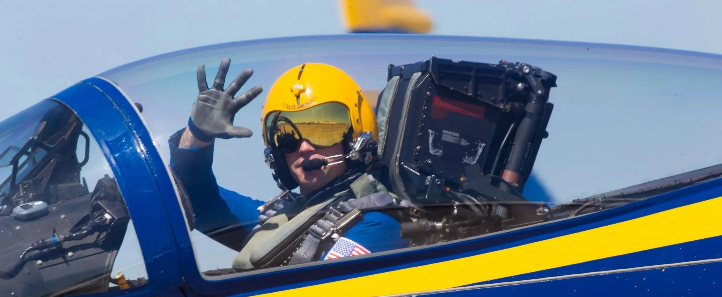 A pilot in a blue flight suit and yellow helmet inside a blue and yellow fighter jet, making an 'OK' gesture with his hand and waving, with a clear sky in the background.
