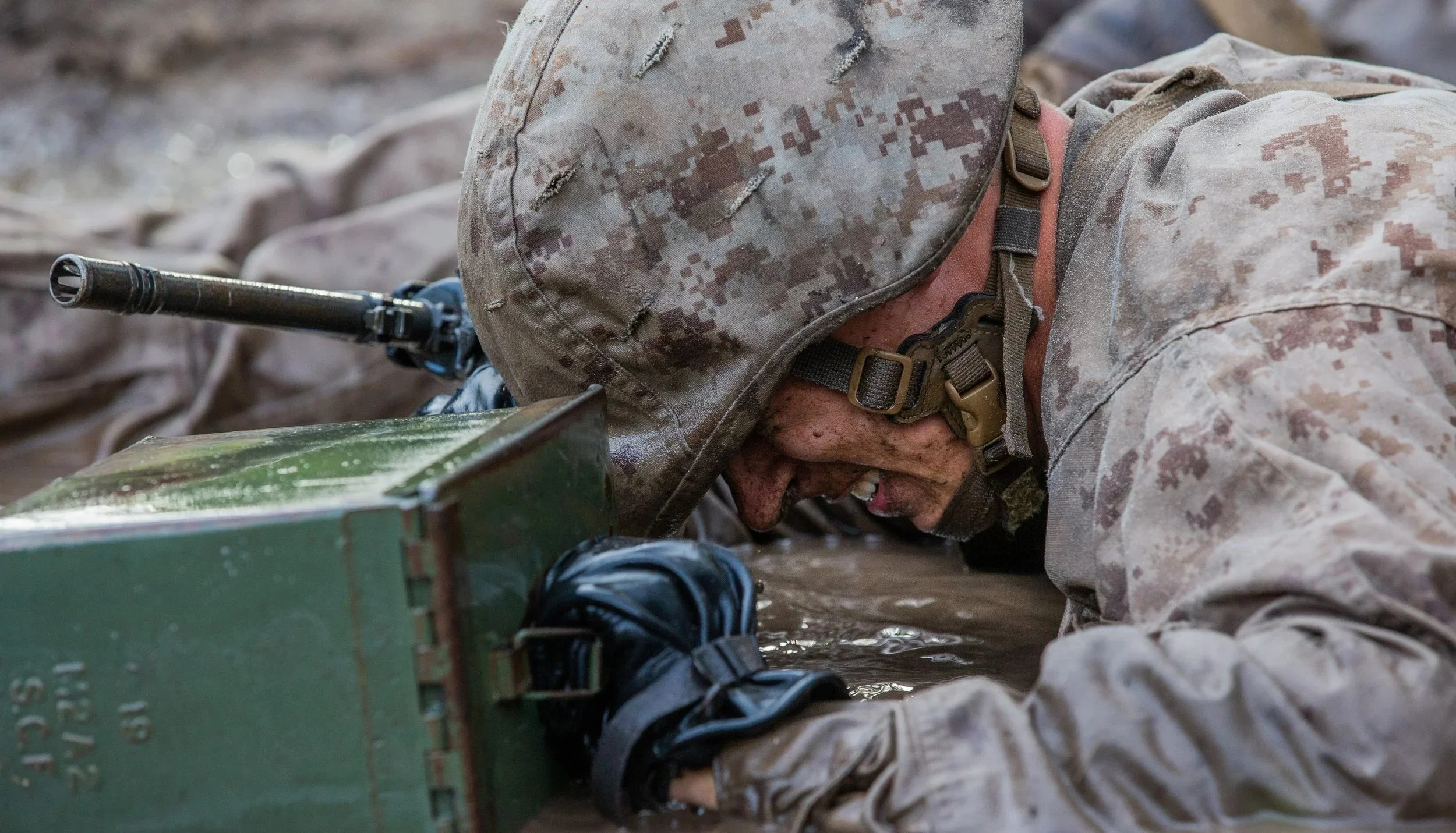 A soldier in camouflage uniform lying on the ground, operating a piece of military equipment with a barrel sticking out, in a muddy environment.
