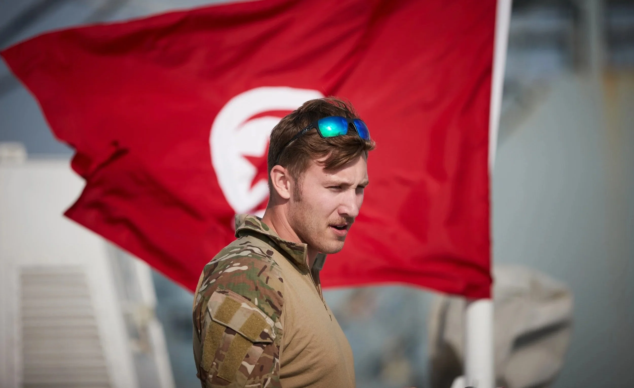A soldier with sunglasses and camouflage uniform standing in front of a large red flag with a white star and crescent symbol.