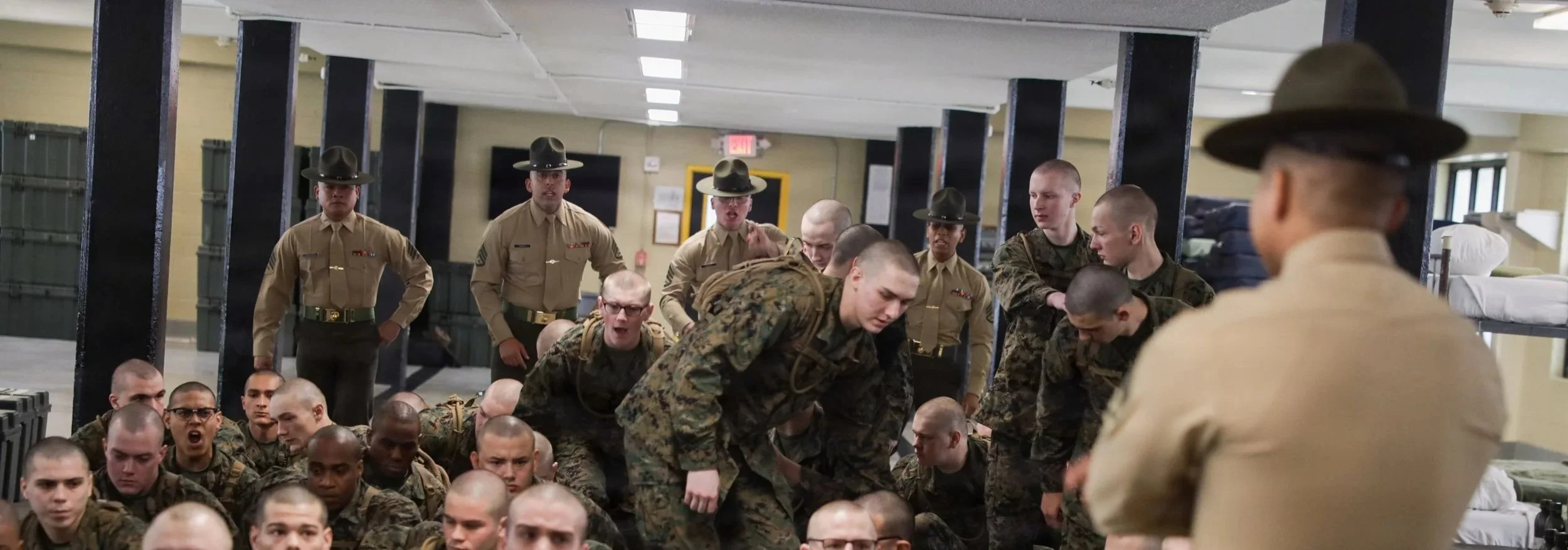 Military recruits in uniform participating in a training exercise in a barracks, with some standing and others kneeling or sitting on the floor.