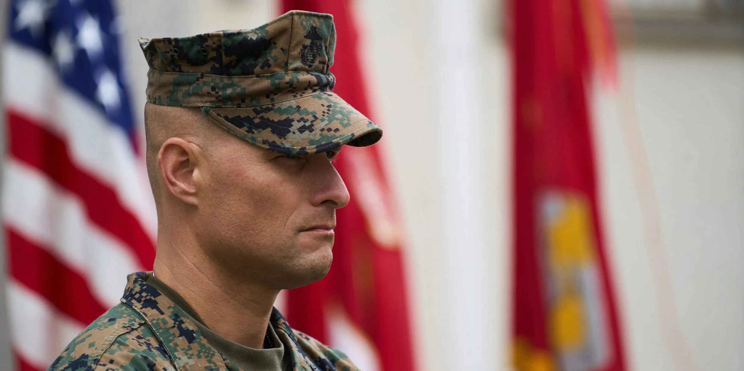 Close-up of a soldier in camouflage uniform, wearing a military cap, with American flags blurred in the background.
