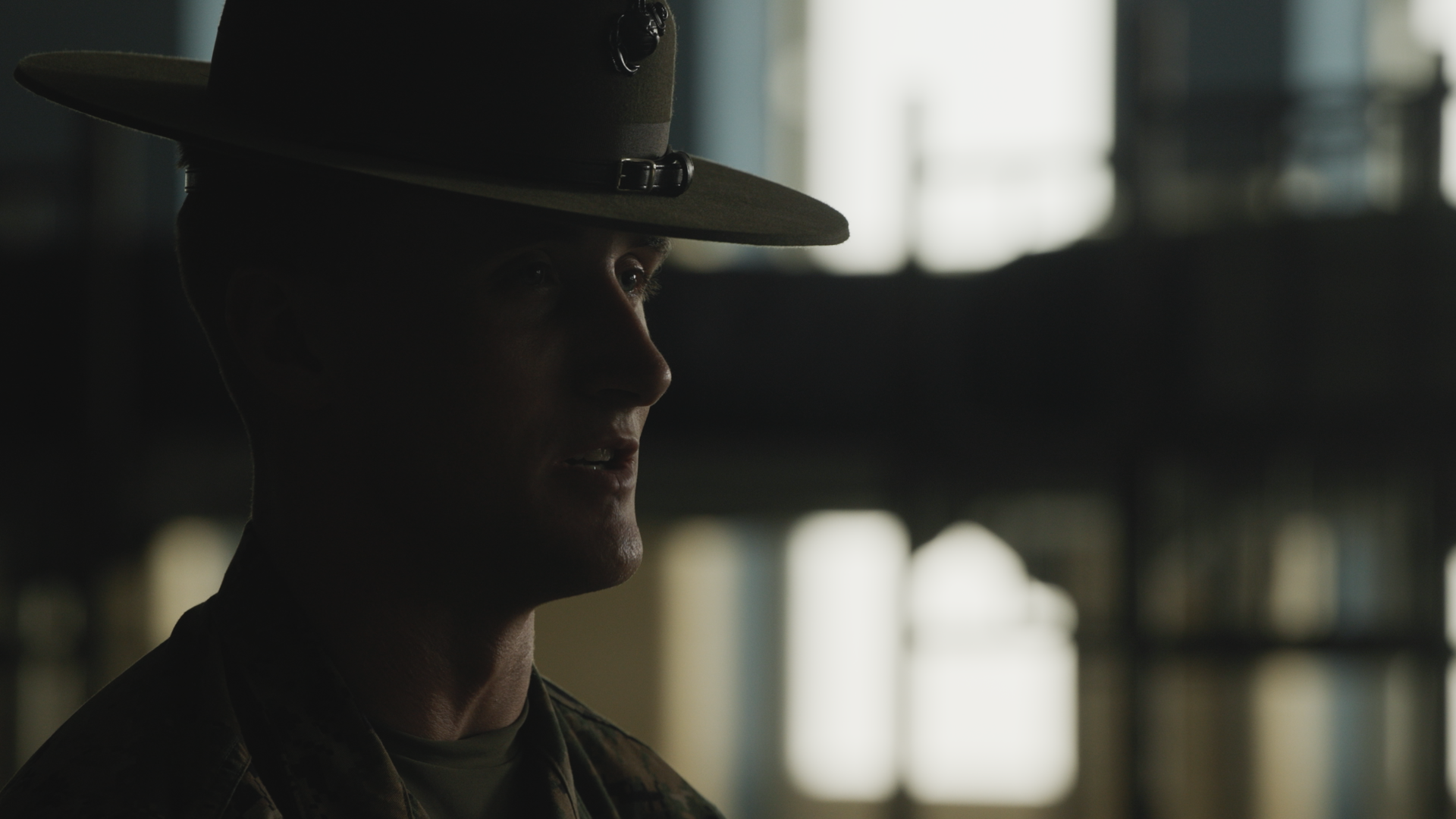Side profile of a young man in military uniform and a drill instructor campaign cover, sitting in a dimly lit room with light coming through windows in the background.