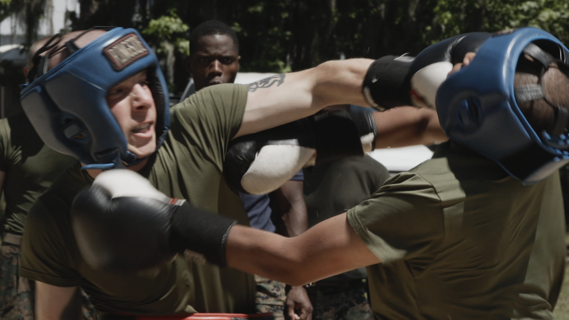 Men dressed in military clothing engage in a physical fight, with one man wearing boxing gloves and a helmet, while another man fights back, in an outdoor setting with trees in the background.