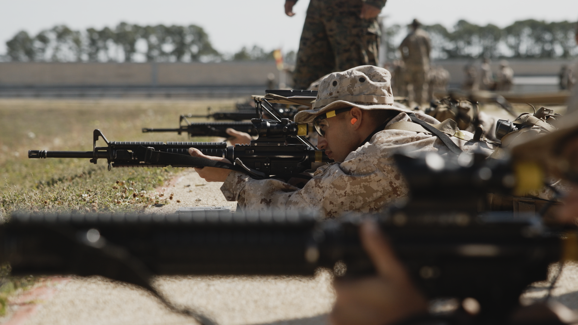 Marines in camouflage uniforms lying prone on the ground and aiming rifles during military training or practice.
