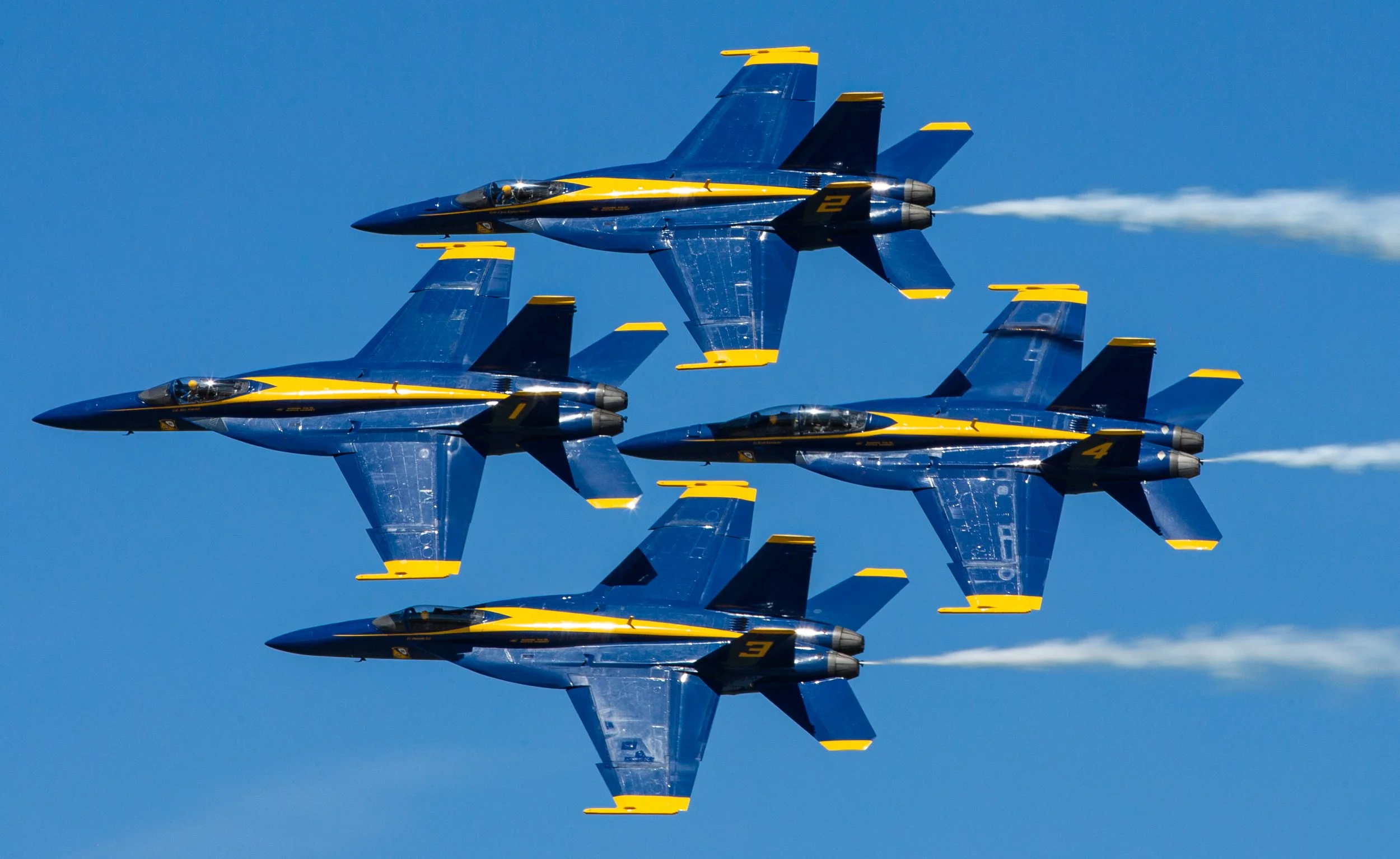 Four blue and yellow fighter jets flying in formation against a clear blue sky, leaving white smoke trails.