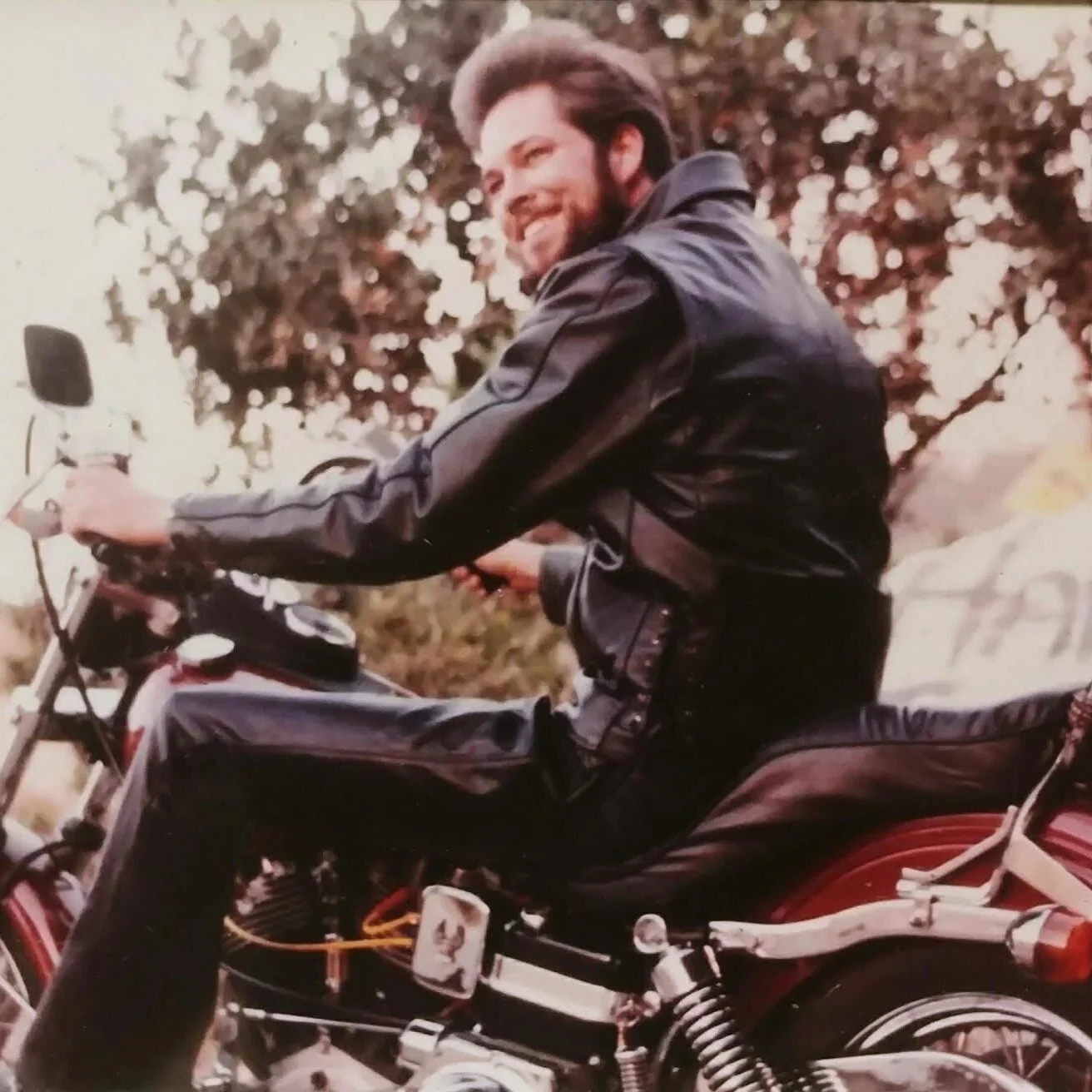 Uncle Bruce, riding a motorcycle outdoors, smiling and wearing a black leather jacket, with trees in the background.