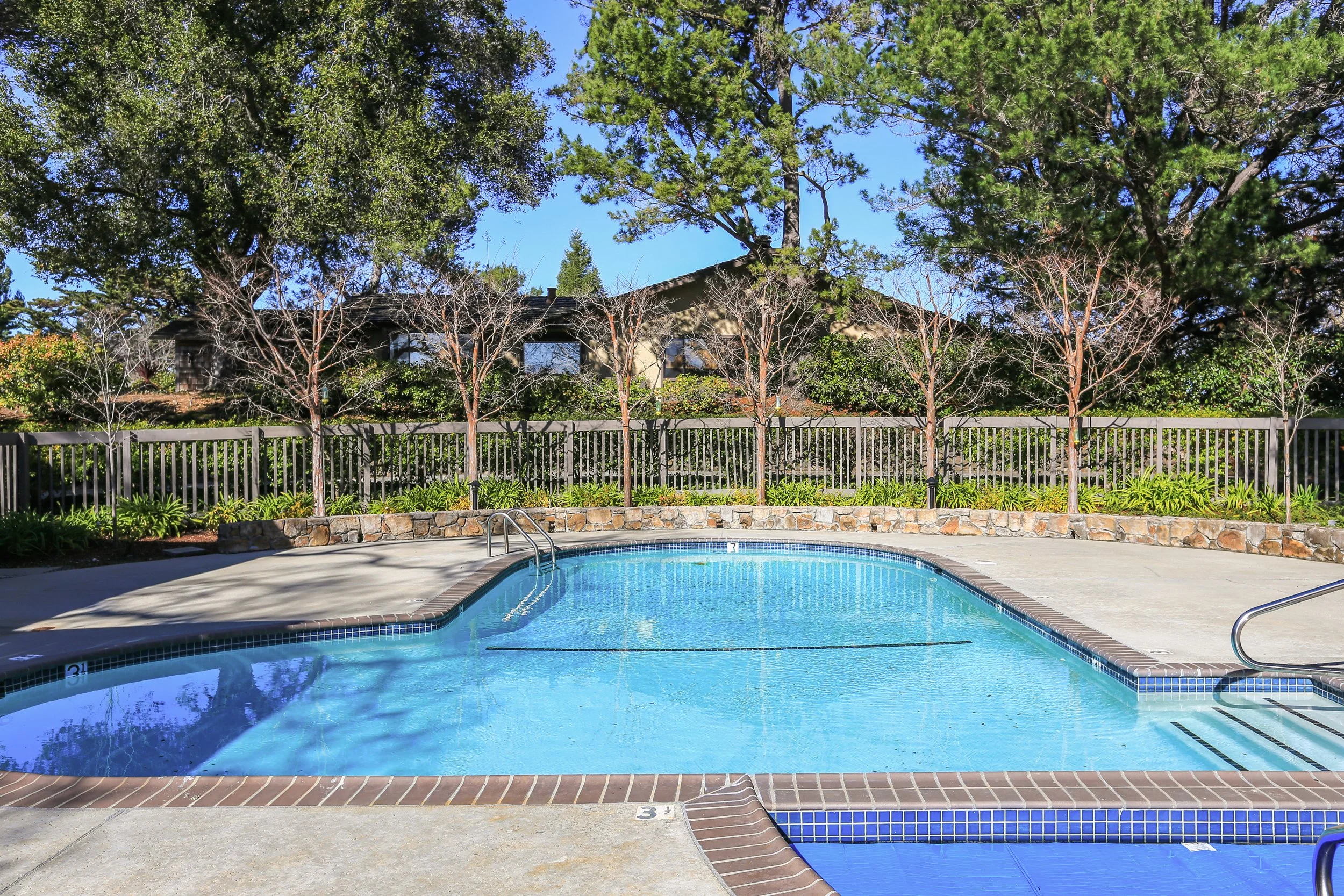 An outdoor swimming pool surrounded by a concrete deck and a metal fence, with trees and a house in the background under a clear blue sky.