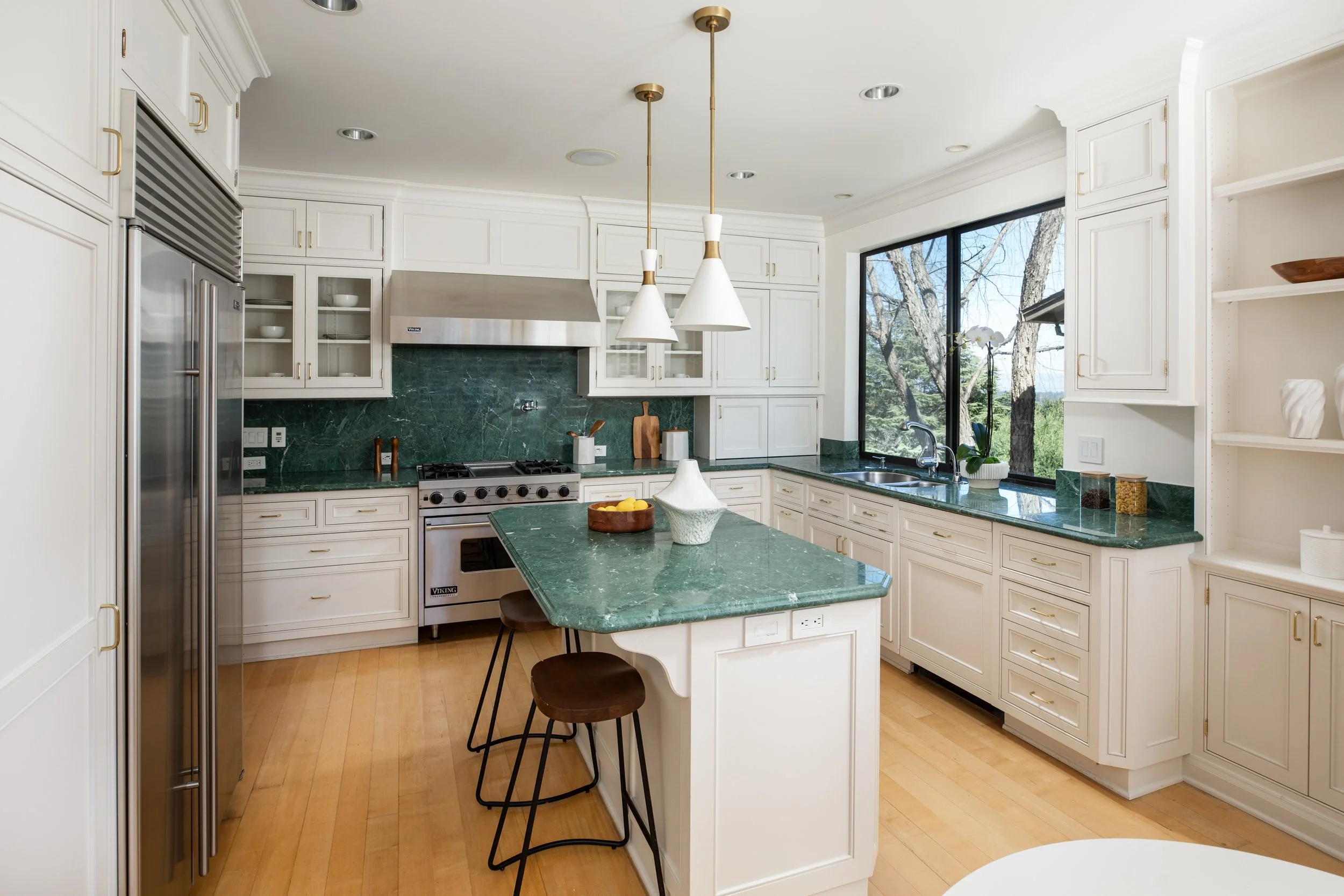 Bright kitchen with white cabinets, green marble countertops, and a large window showing trees outside. Features include built-in appliances, pendant lights, some decorative items, and a small breakfast bar with three brown stools.