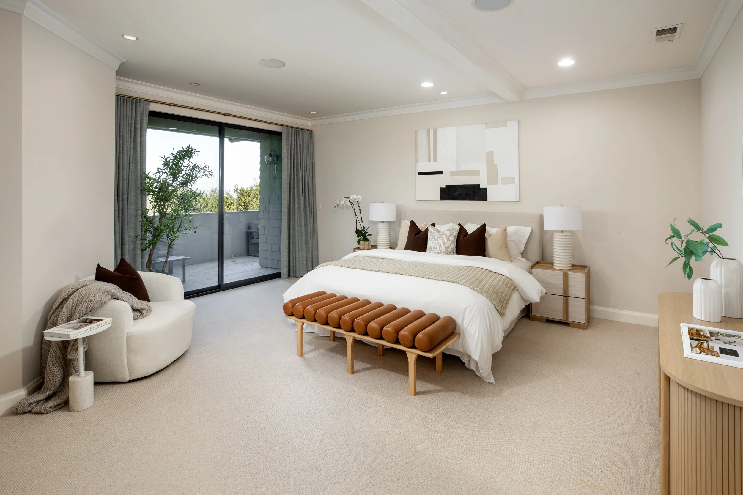 A bedroom with a large bed, nightstands with lamps, a bench at the foot of the bed, a white armchair with a blanket and book, a sliding glass door with curtains leading to a balcony, and a large abstract painting on the wall.