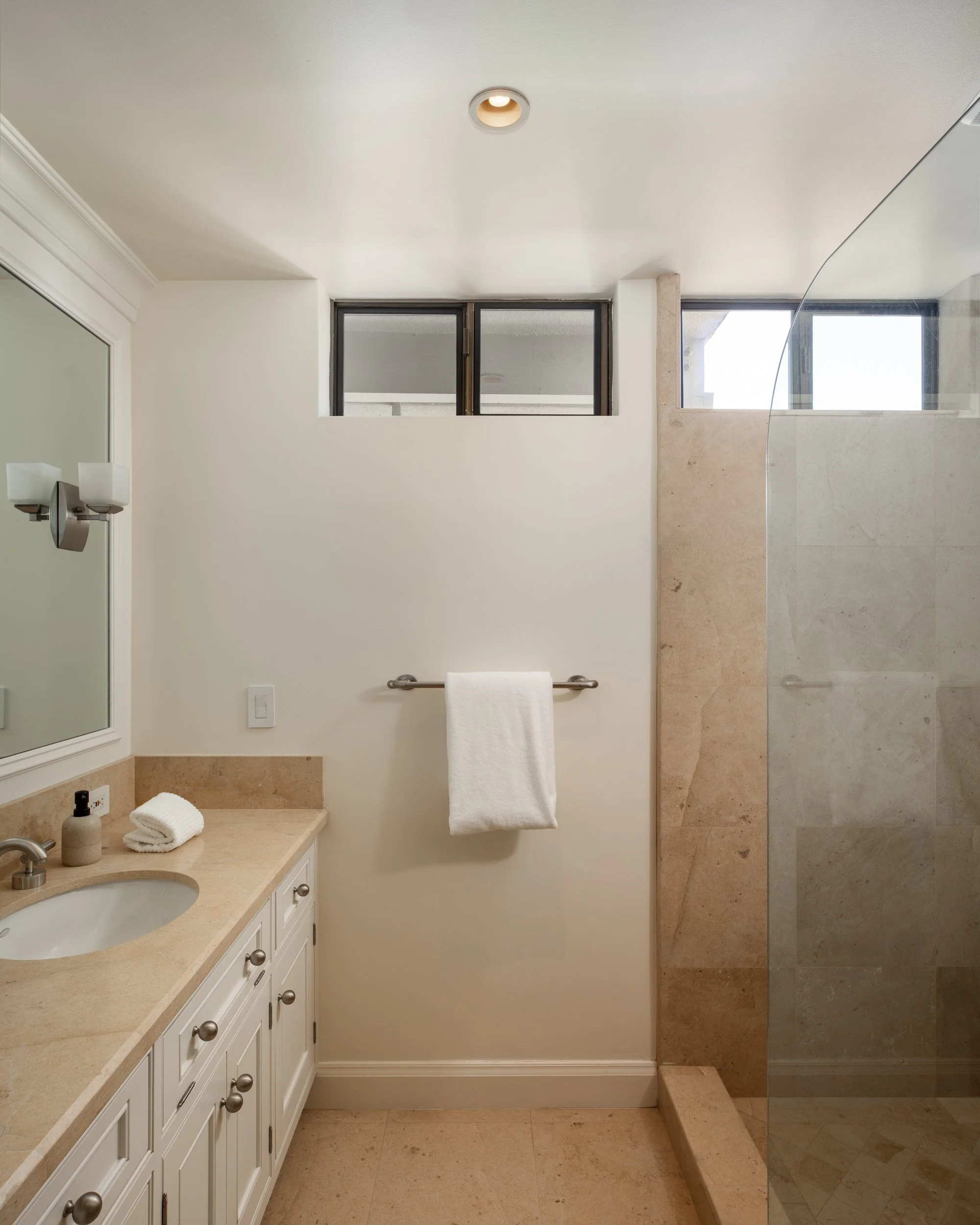 A bathroom with a vanity on the left side, featuring a beige countertop, a sink, and a mirror with a wall-mounted light fixture. Towels are hanging on a bar, and there's a soap dispenser and rolled towel on the countertop. Behind the bathroom, there'