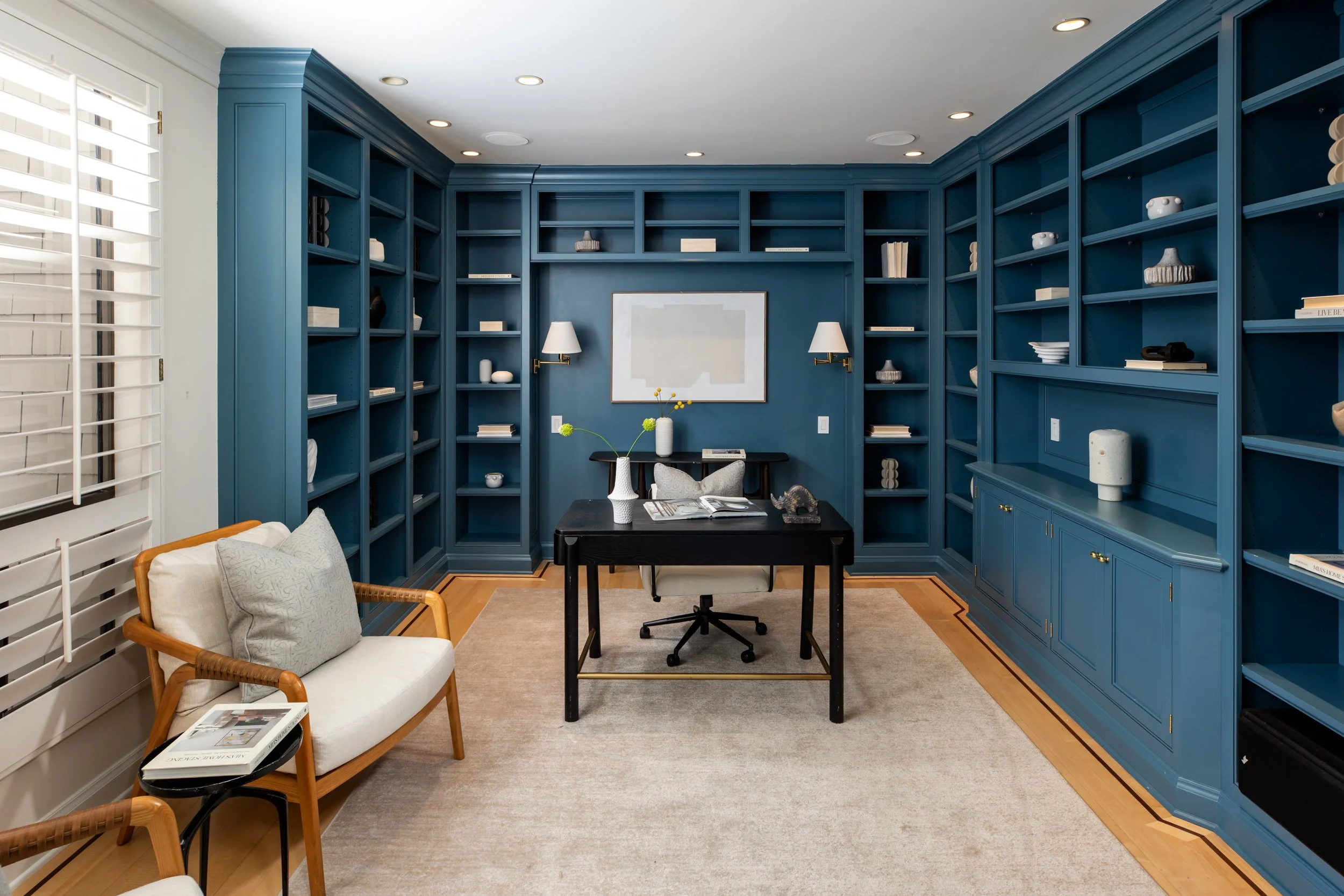 A home office with built-in blue bookshelves, a black desk, an ergonomic chair, a beige armchair with a pillow, and wall-mounted lamps.
