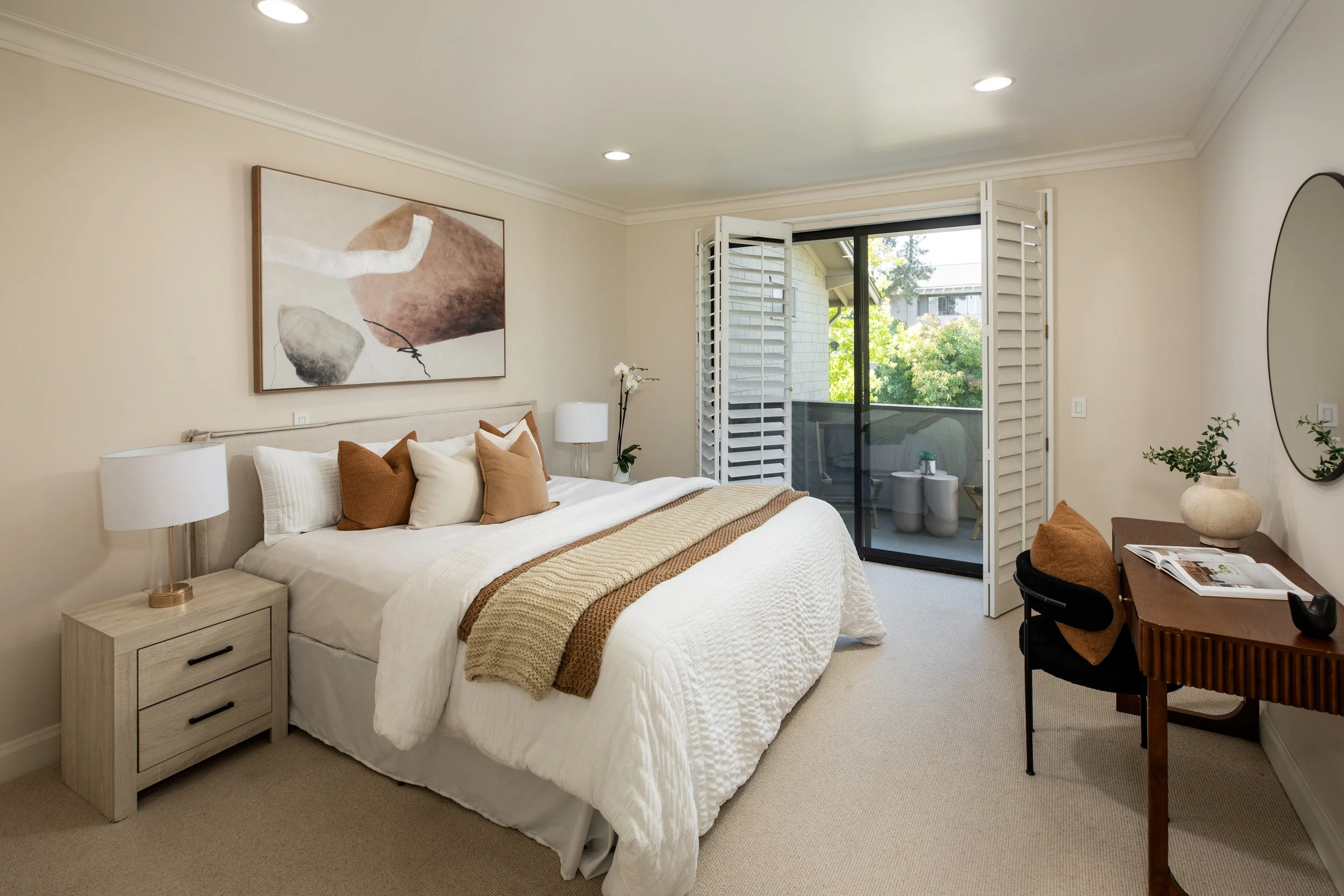 A modern bedroom with a large bed decorated with white and beige pillows, a beige throw blanket, and a wooden nightstand with a lamp. There is a abstract painting above the bed, a balcony with open white shutters, and a small sitting area with a roun