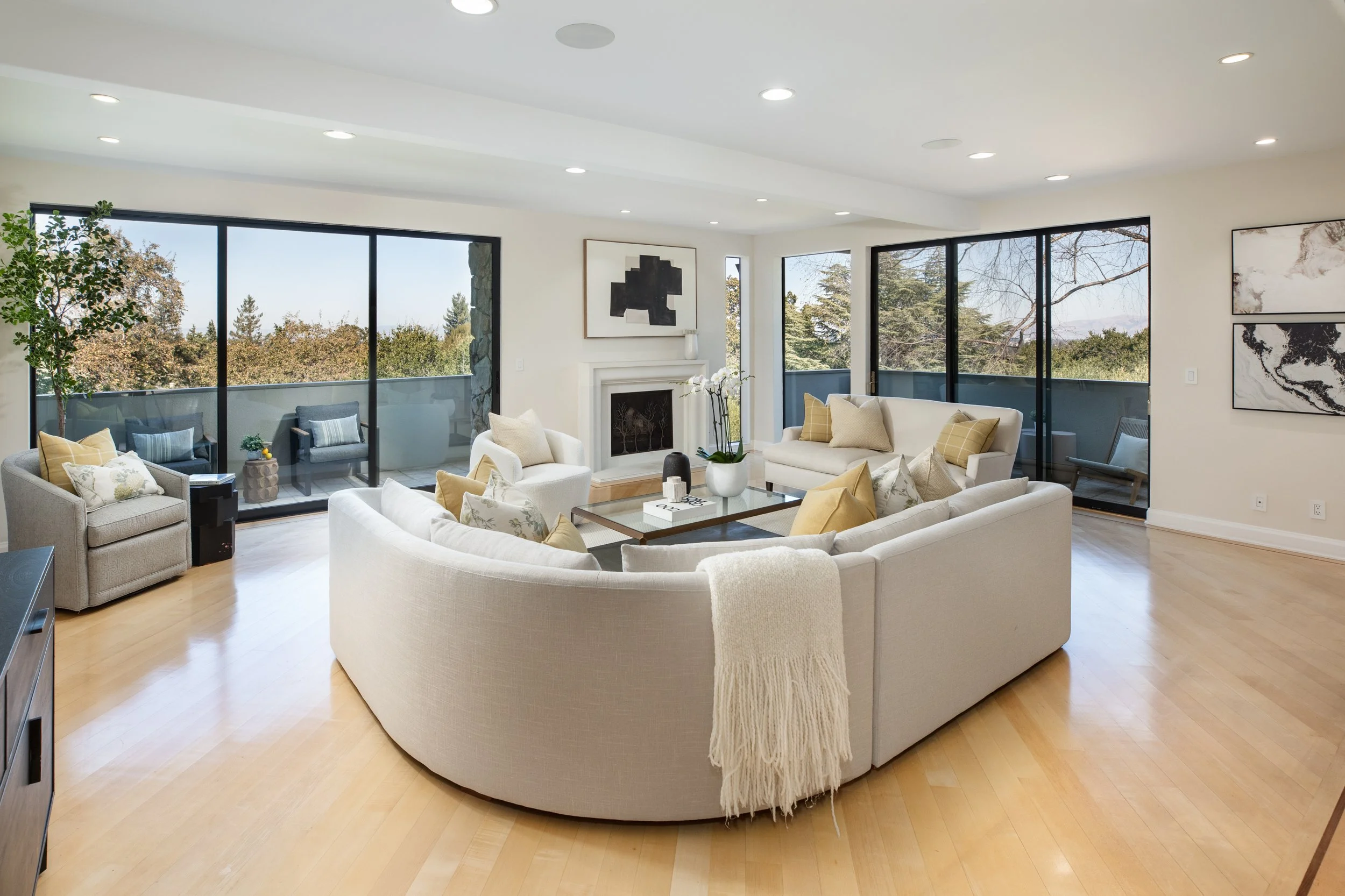 Bright living room with large windows showing trees outside, furnished with white sofas, armchair, coffee table, and wall art.