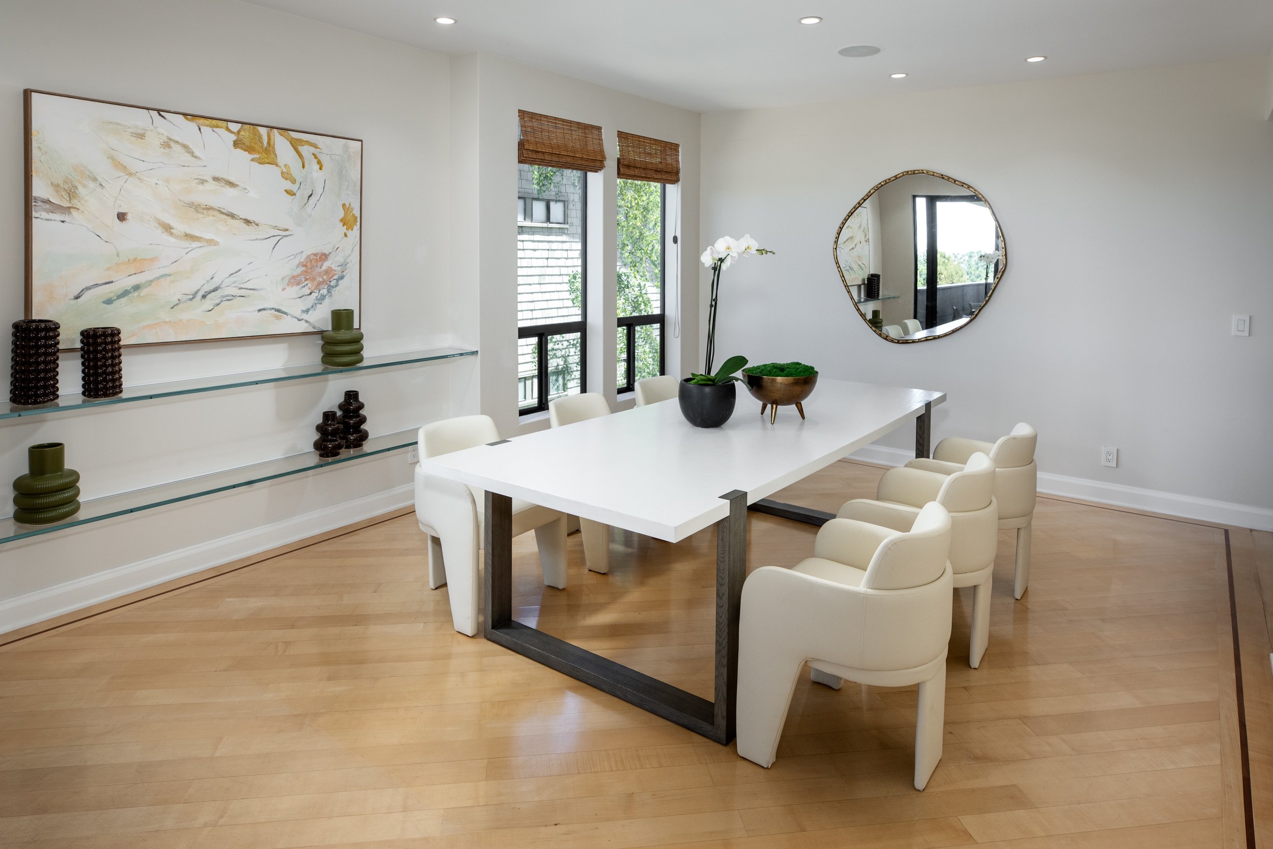 A modern dining room with a white rectangular table surrounded by six beige chairs. The room has three windows with bamboo blinds, a large abstract painting on the wall, and a round mirror. Decor includes vases, a potted orchid, and a bowl of moss on