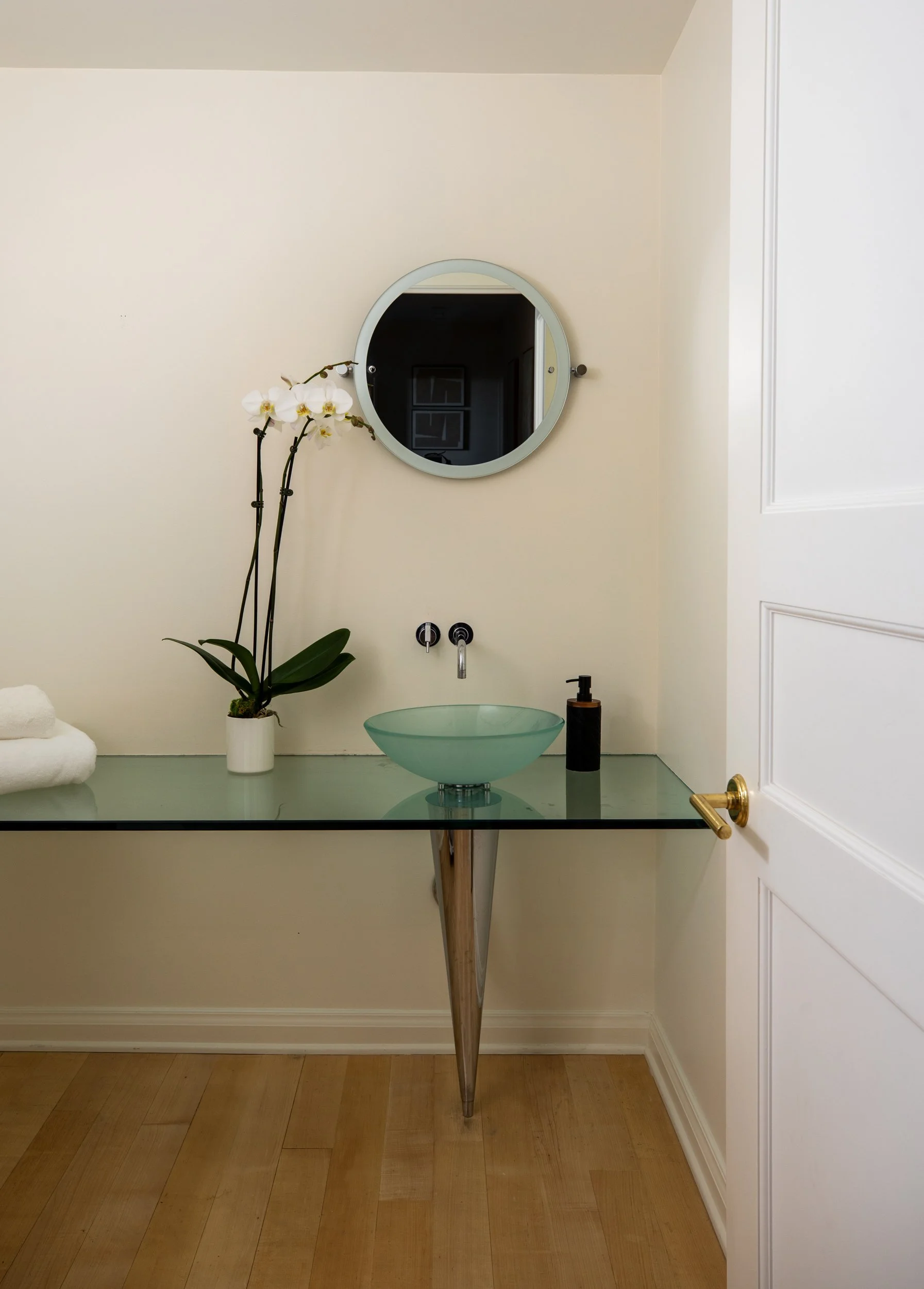 A bathroom countertop with a glass vessel sink, a soap dispenser, a white towel, a potted orchid, a round mirror, and a white cabinet door on the right.