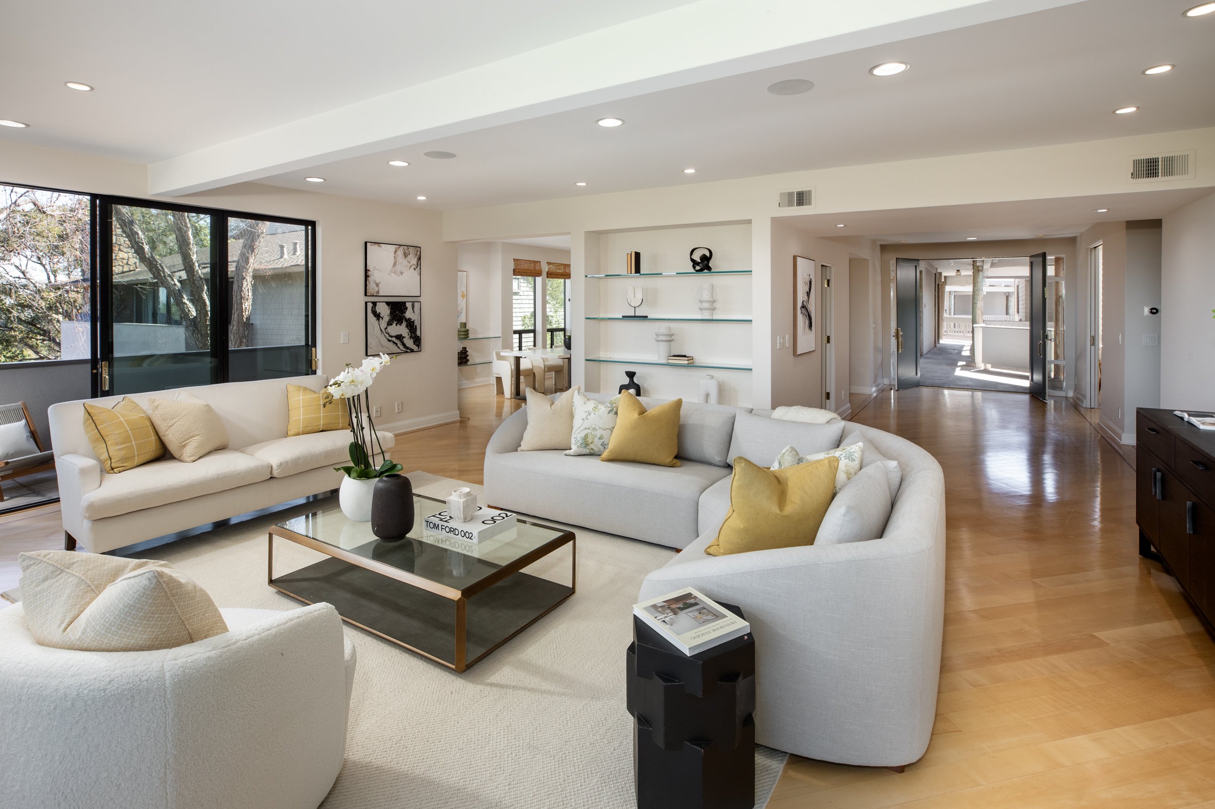 Modern living room with white sofas, yellow pillows, glass coffee table, and large windows showing outdoor trees.