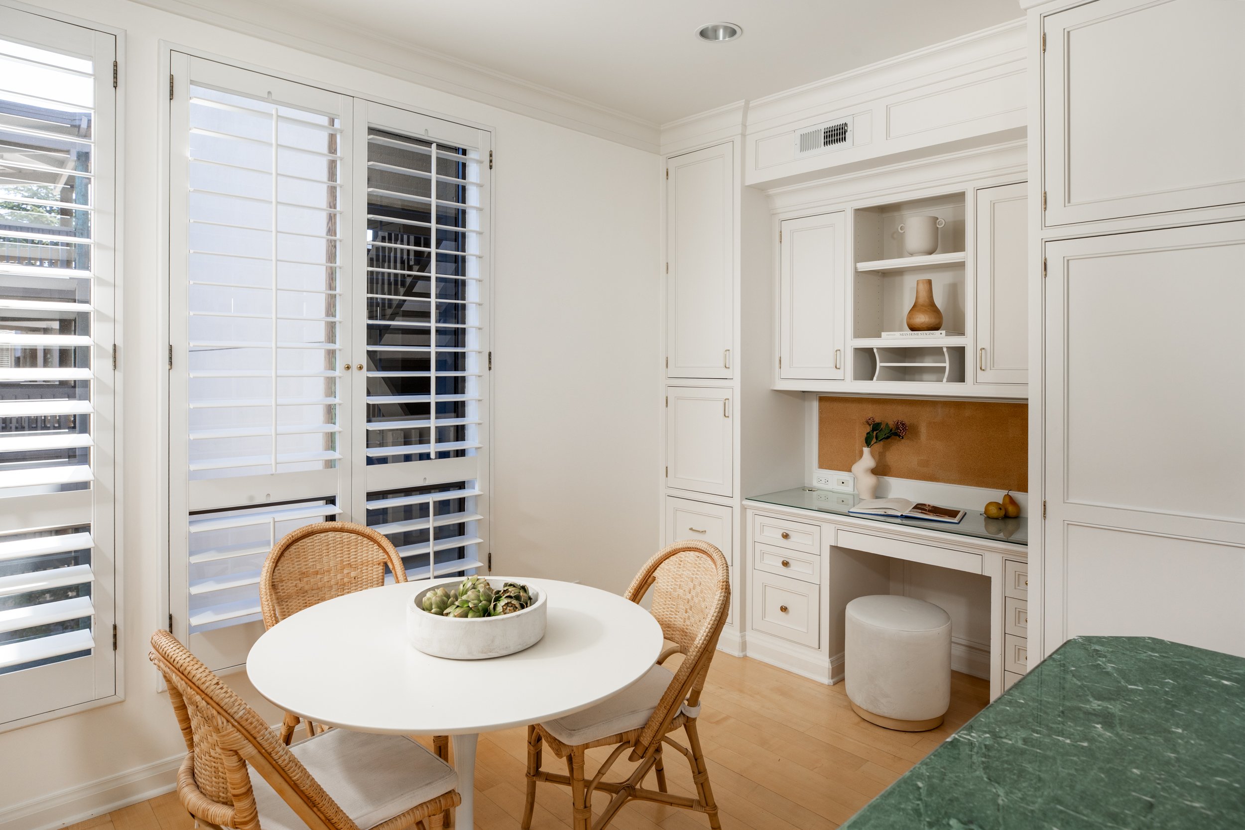 White dining room with a round table, wicker chairs, built-in cabinets, and a window with white shutters.
