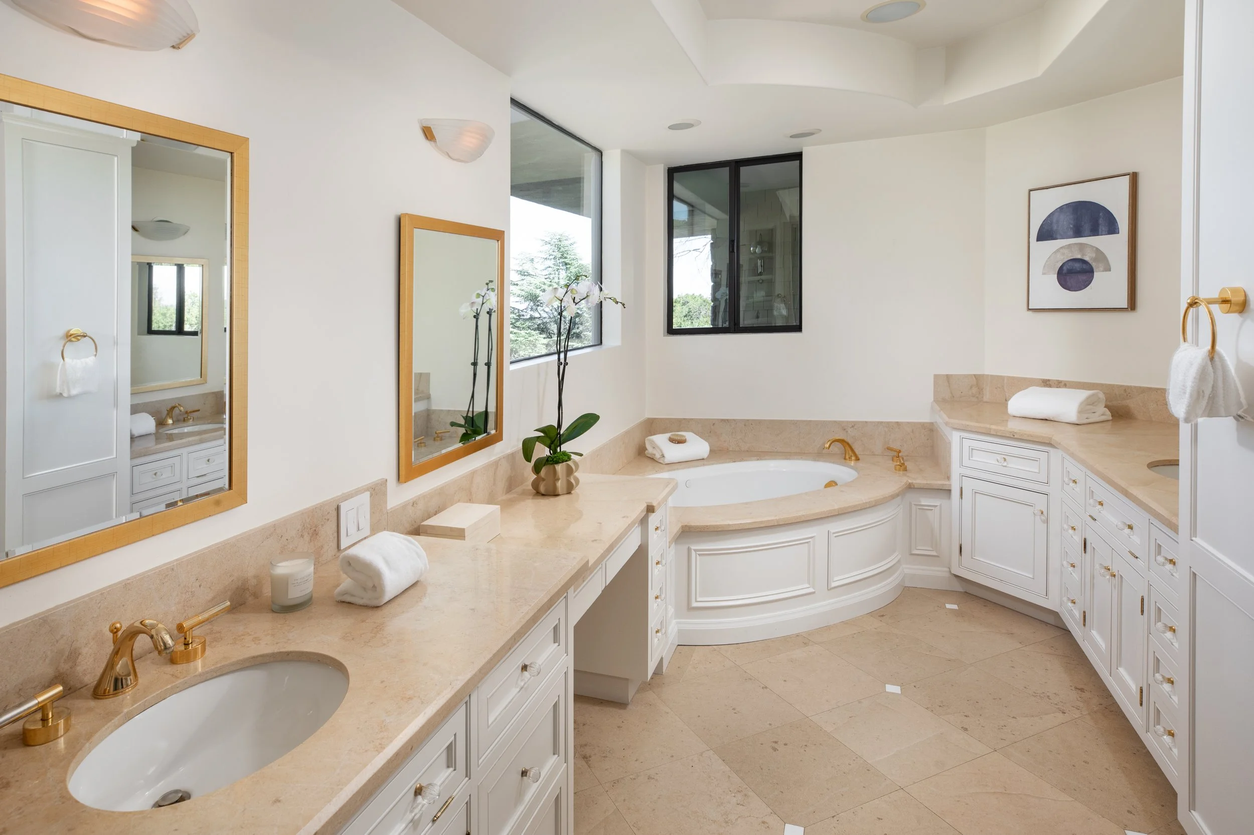 Bright bathroom with white cabinetry, beige marble countertops, and a large corner bathtub. There are two mirrors with gold frames above the sinks, and decorative vases with orchids. The room has windows that let in natural light and minimal wall art