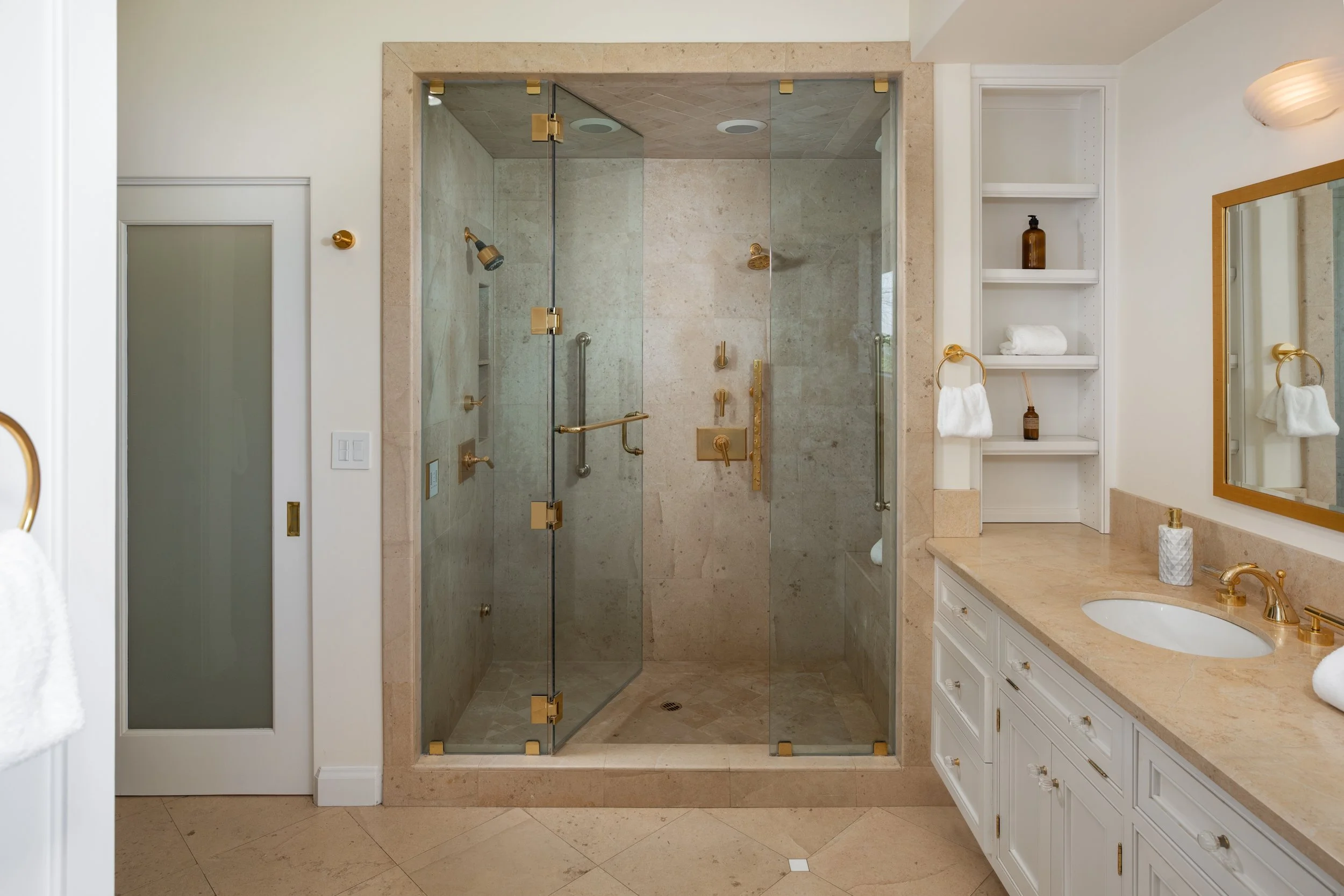 Modern bathroom with a glass-enclosed shower featuring brass hardware, a white vanity with a beige marble countertop, and built-in shelves with toiletries and towels.
